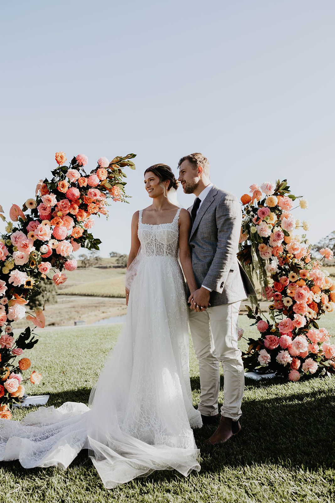 Un couple se tient devant une arche ornée de fleurs, avec un champ verdoyant en arrière-plan.
