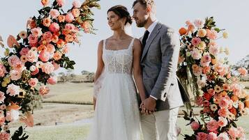 Un couple se tient devant une arche ornée de fleurs, avec un champ verdoyant en arrière-plan.
