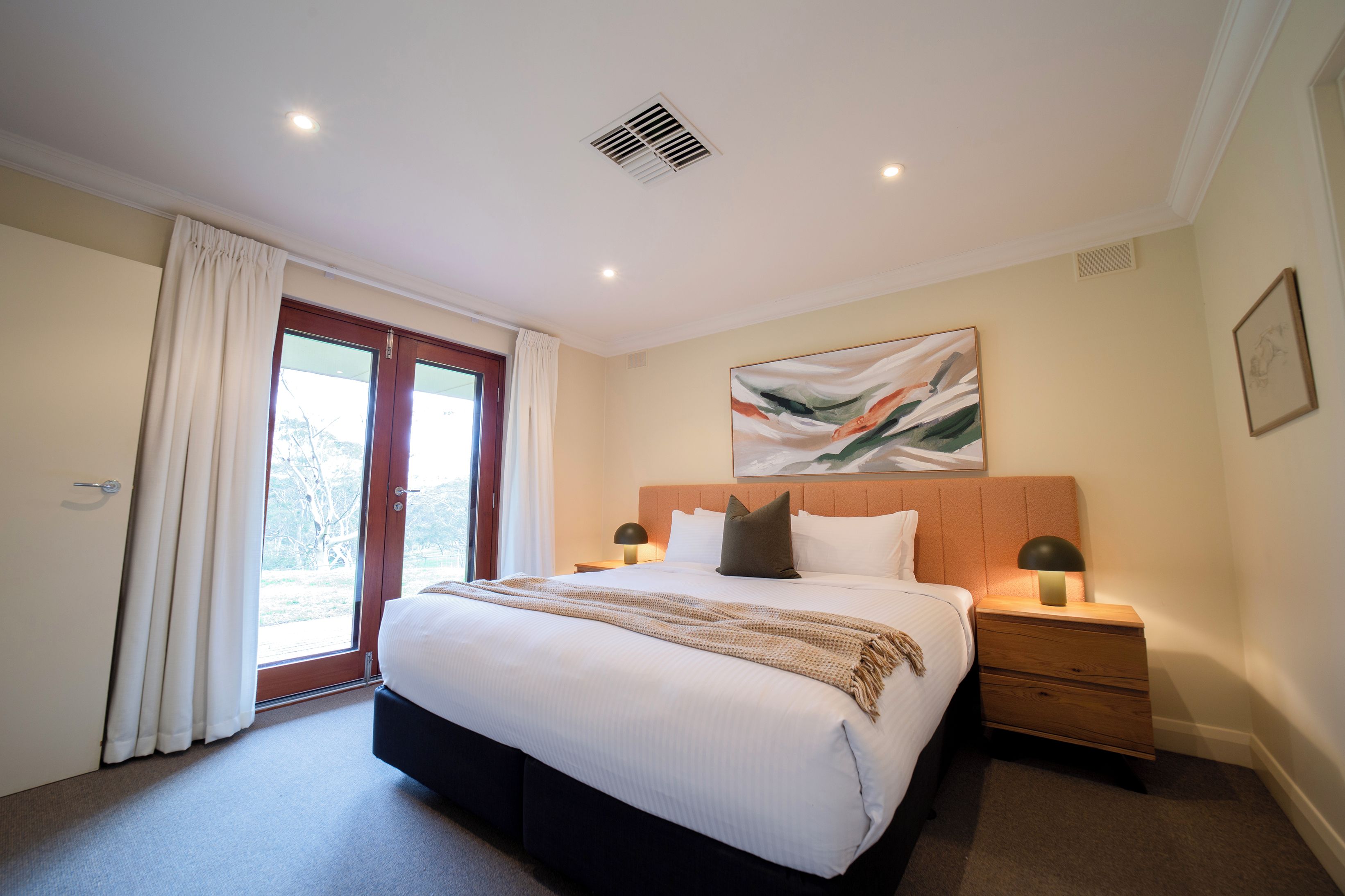 Una habitación de hotel con una cama grande, dos mesitas de noche con lámparas, una ventana con cortinas y una rejilla en el techo.