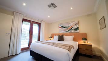 Una habitación de hotel con una cama grande, dos mesitas de noche con lámparas, una ventana con cortinas y una rejilla en el techo.