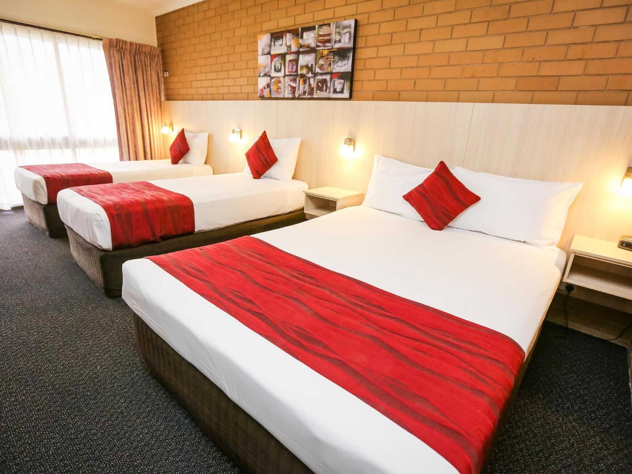 Une chambre d’hôtel avec trois lits, chacun recouvert d’une couverture rouge et blanche assortie aux oreillers. Un mur de briques est décoré d’un tableau encadré placé au-dessus des lits.