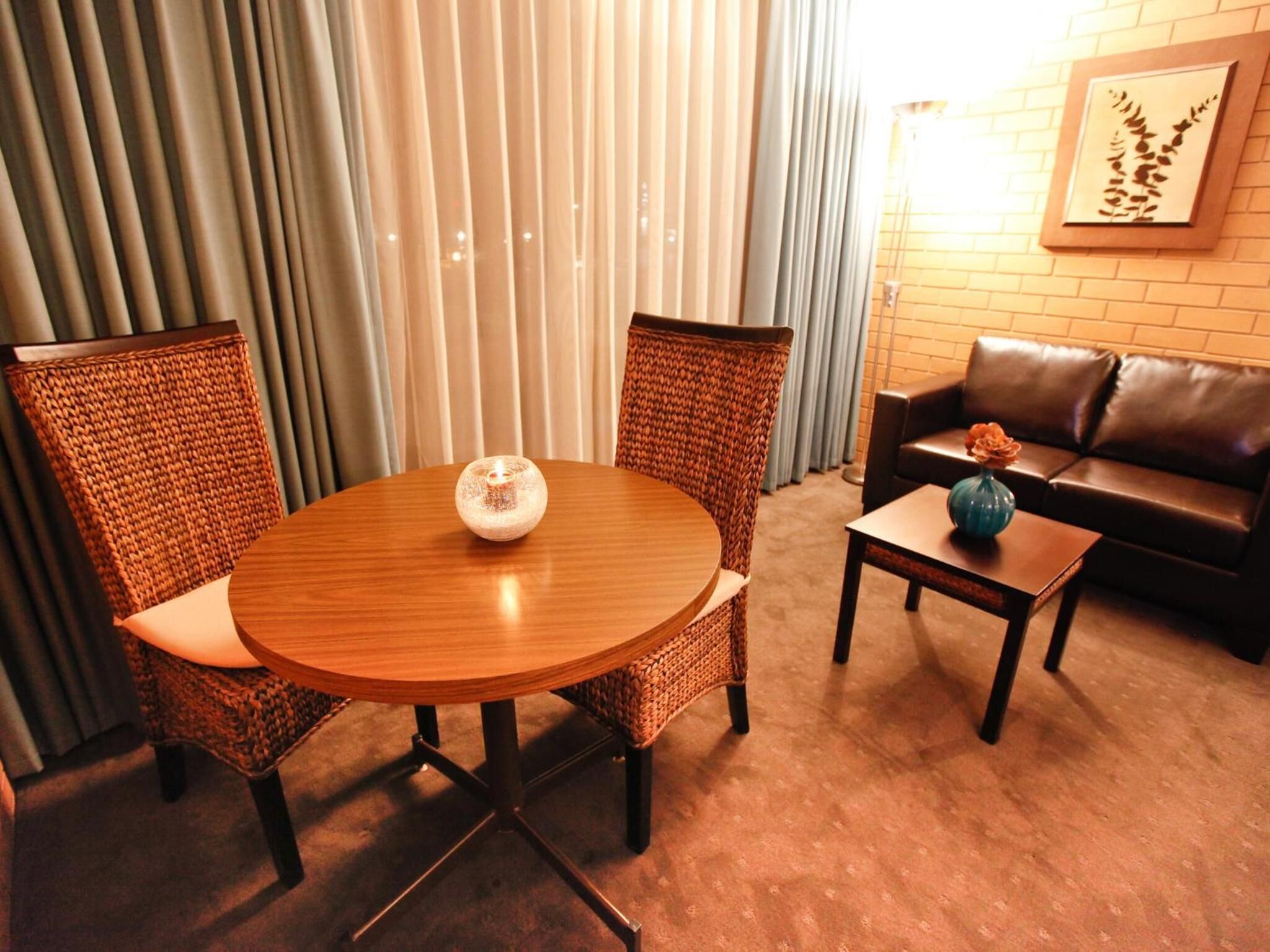 A round wooden table with a candle, two wicker chairs, a small table with a vase, and a leather sofa in a warmly lit room with vertical blinds.