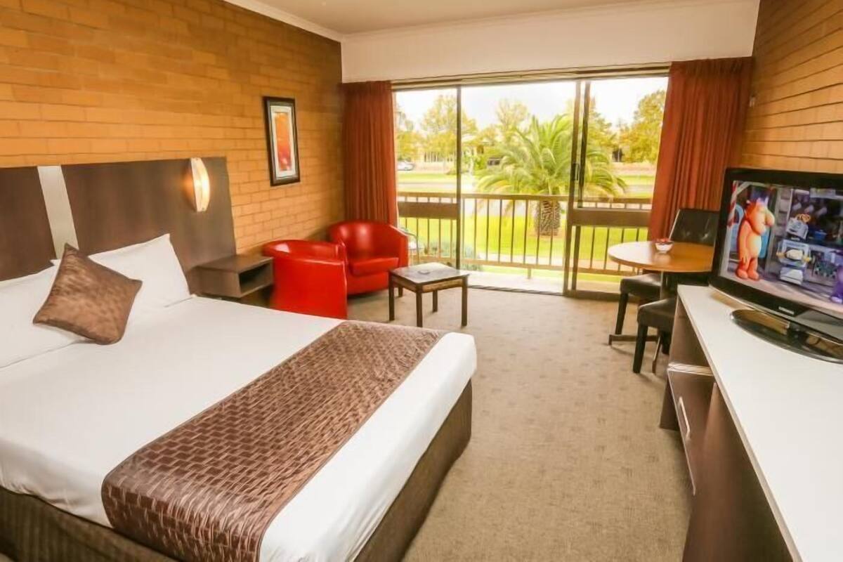 A hotel room with a large bed, a red chair, a small table, a TV, and a balcony with a view of palm trees.