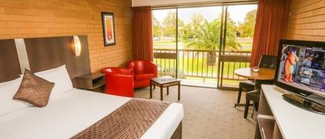 A hotel room with a large bed, a red chair, a small table, a TV, and a balcony with a view of palm trees.