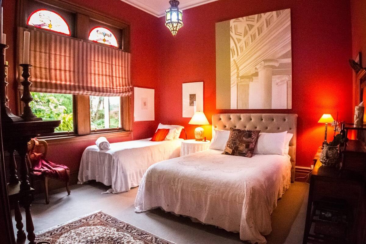 A hotel room with two beds, a large window with curtains, a chandelier, and a painting on the wall.