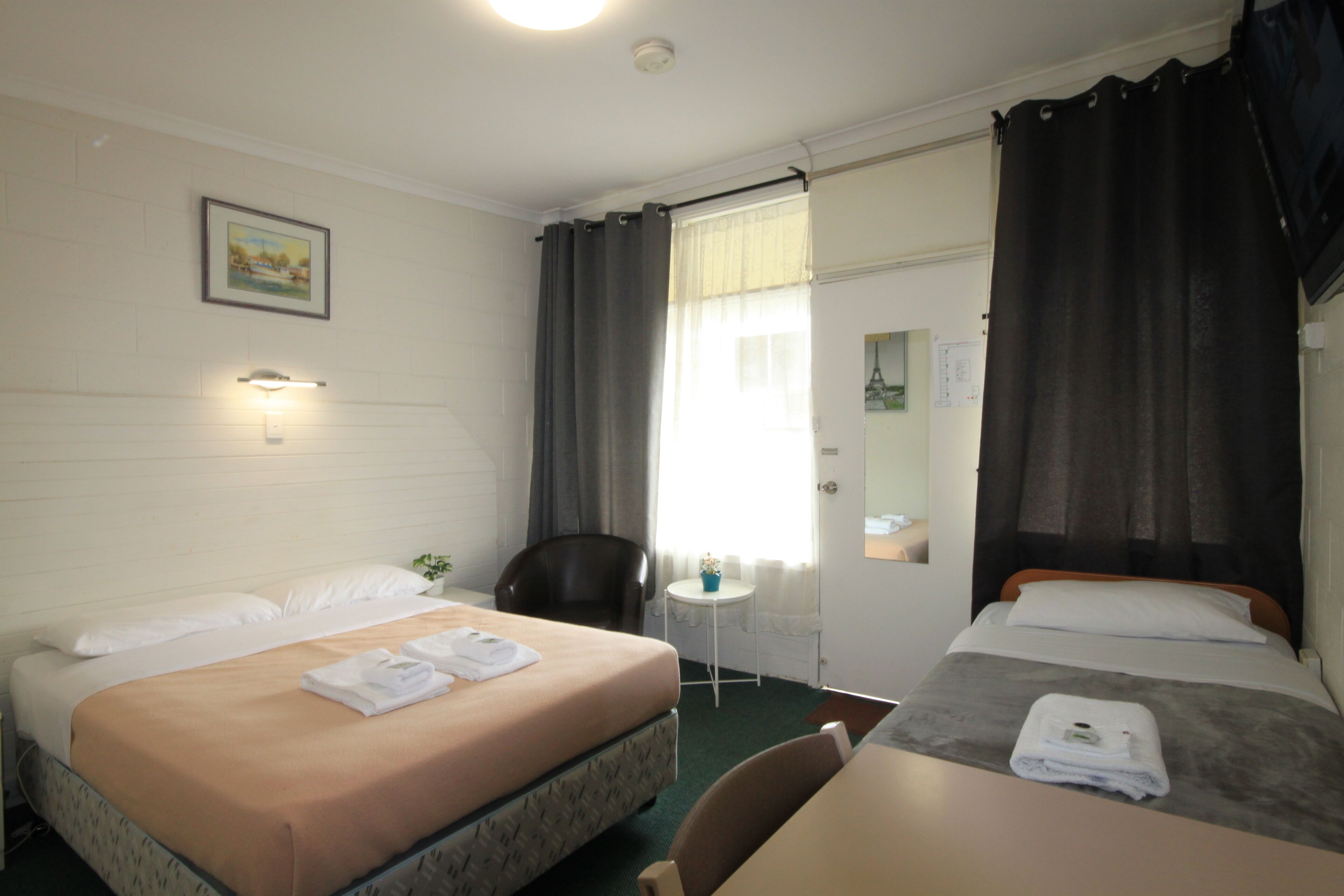 Une chambre d’hôtel avec deux lits, une petite table, une chaise, un bureau, un miroir et une fenêtre avec des rideaux.