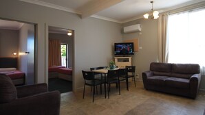 A living room with a sofa, a dining table with chairs, a television, and a window with curtains. - Pelicans Motel San Remo (San Remo)
