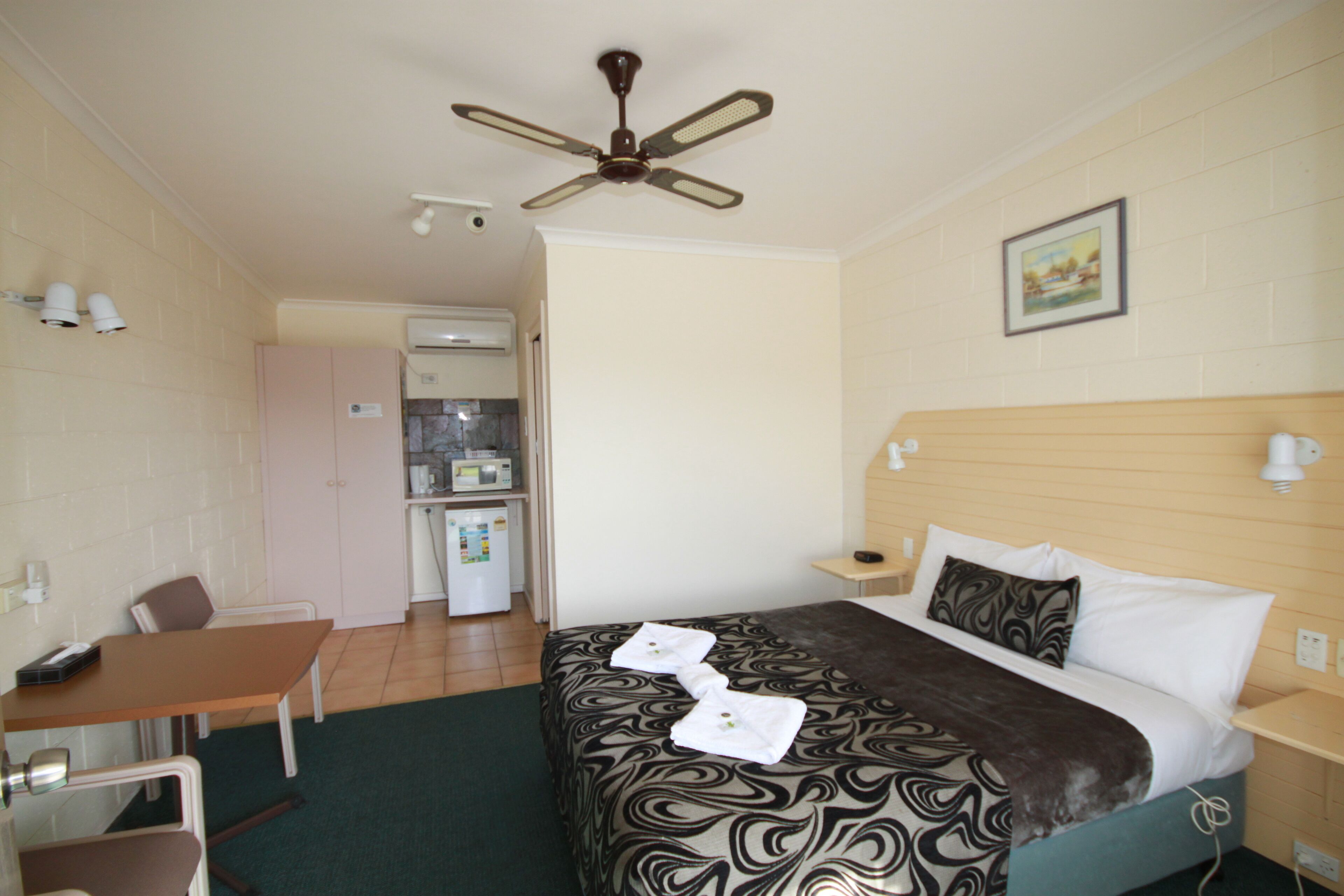 Camera d'albergo con un letto, una scrivania, una sedia, una ventola a soffitto, un quadro appeso al muro e un angolo cottura sullo sfondo.
