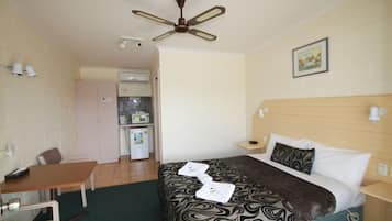 Camera d'albergo con un letto, una scrivania, una sedia, una ventola a soffitto, un quadro appeso al muro e un angolo cottura sullo sfondo.