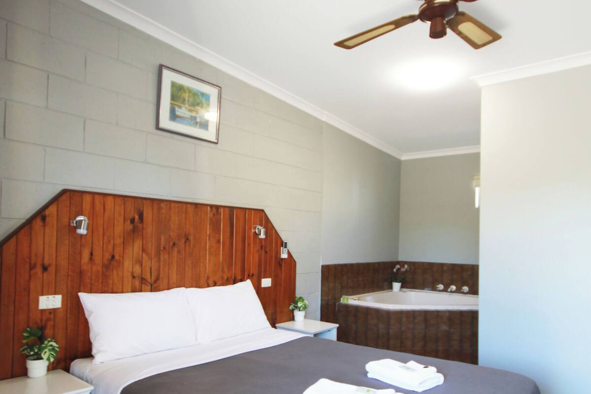 A bedroom with a wooden headboard, a bed with grey bedding, a bedside table with a plant, a nightstand, and a ceiling fan.