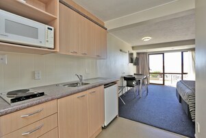 Cocina compacta con microondas, lavaplatos y fregadero, que da a un balcón con zona de comedor y cama.