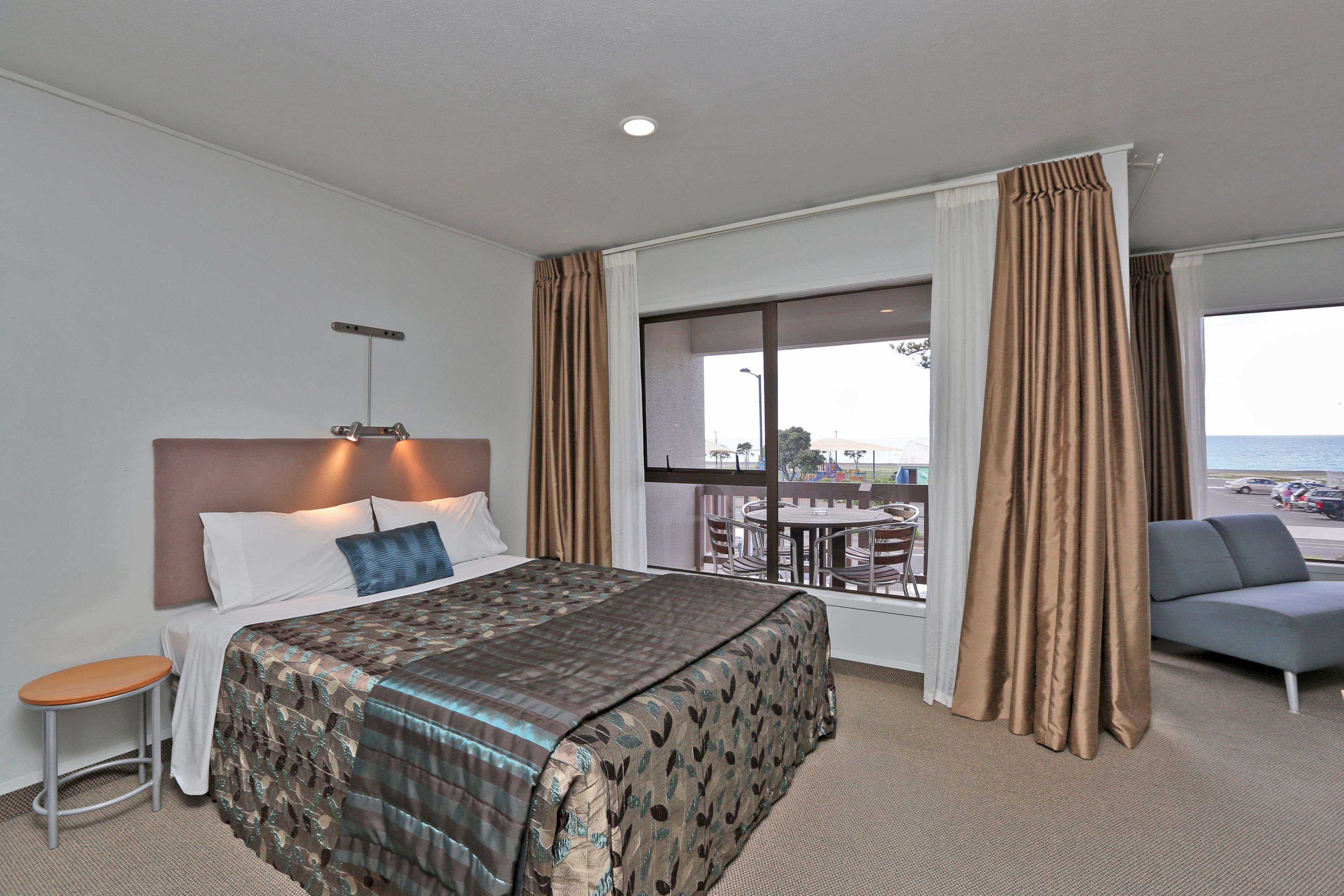 A hotel room with a bed, a small table, a sofa, and a large window with curtains.