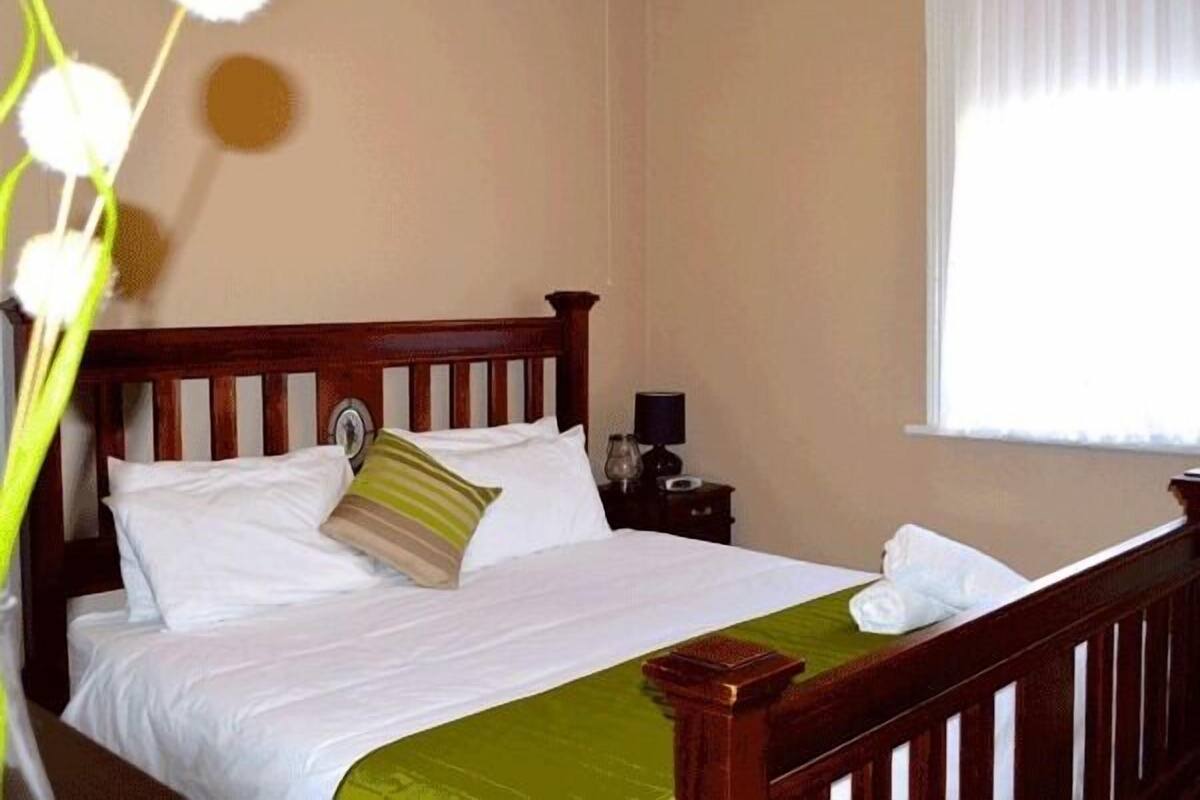 A neatly made bed with white linens and a green bedspread, a wooden headboard, a bedside table with a lamp, and a window with sheer curtains.