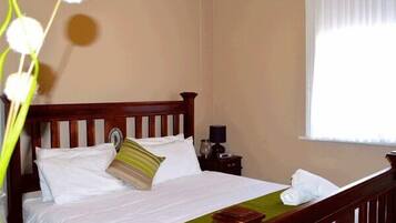 A neatly made bed with white linens and a green bedspread, a wooden headboard, a bedside table with a lamp, and a window with sheer curtains.