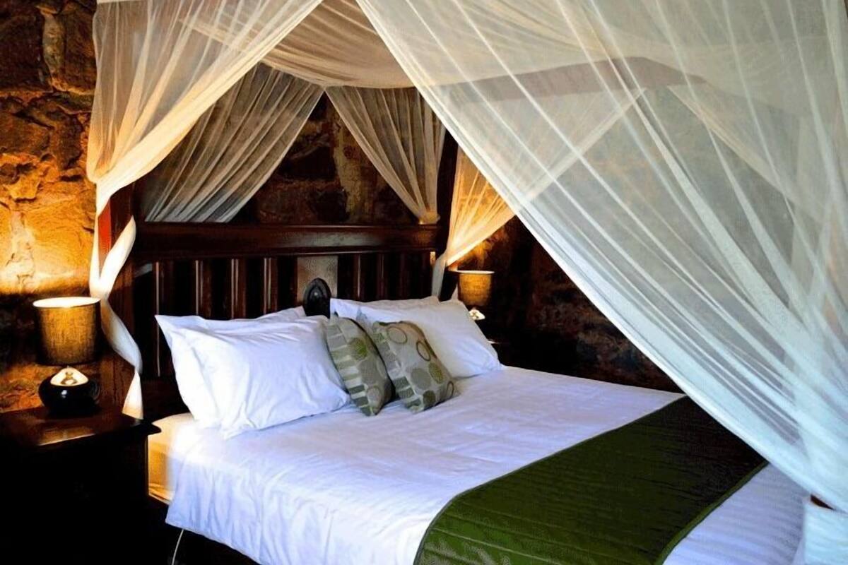 A bed with white linens, a wooden headboard, and a green throw blanket. There is a nightstand with a lamp and a small decorative item. The room has a stone wall background.