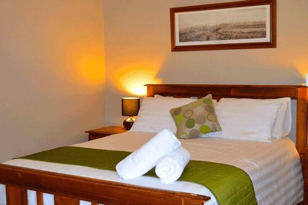 A neatly made bed with a green bedspread, white pillows, and a rolled white towel. There is a framed picture on the wall above the bed.