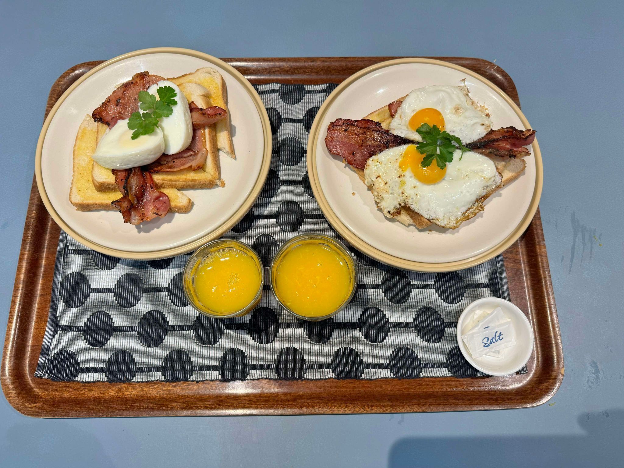 A breakfast tray with two plates of eggs and bacon, two glasses of orange juice, and a small container of salt.