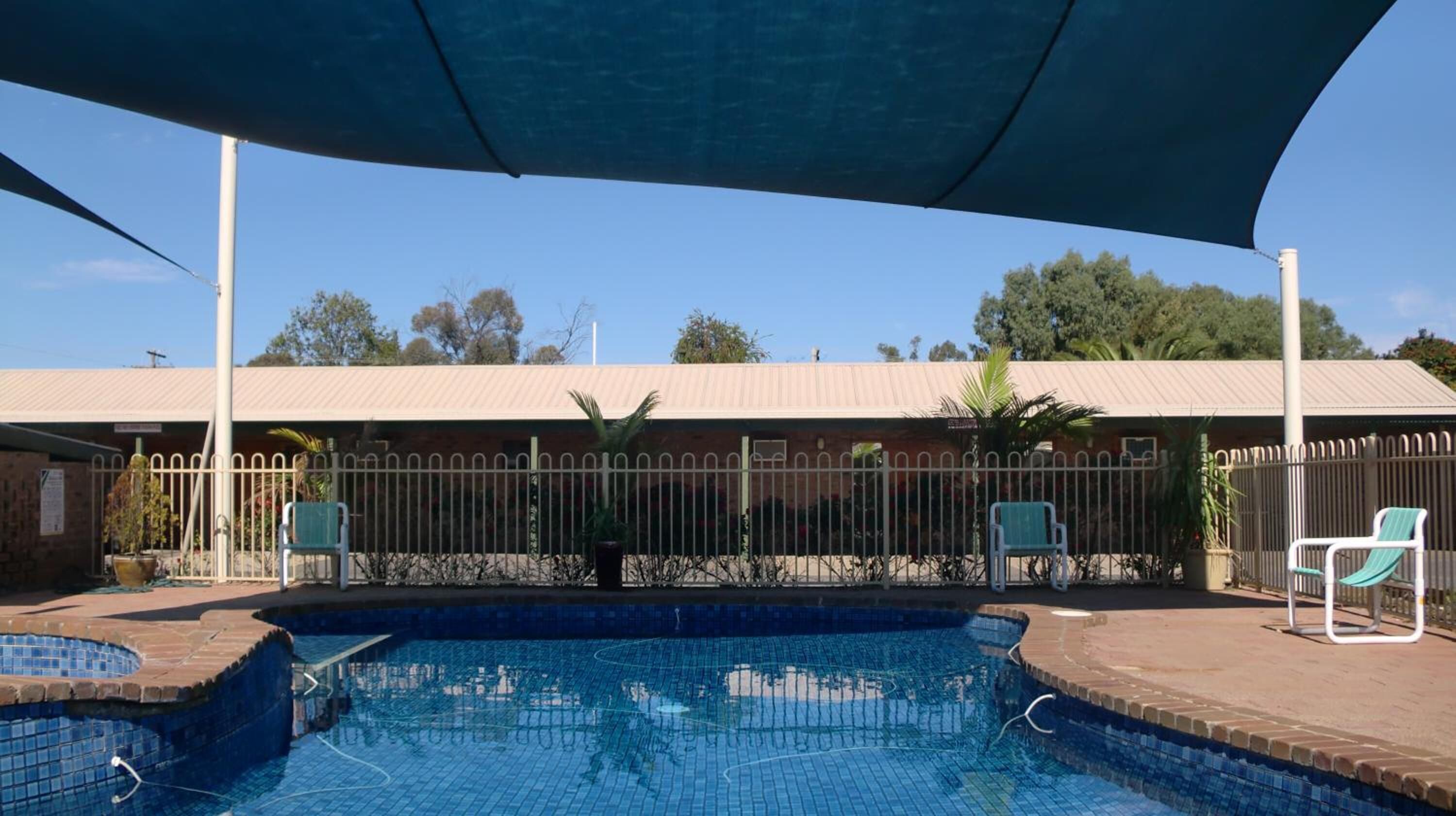 A swimming pool with a shaded area, a fenced enclosure, and a building in the background.