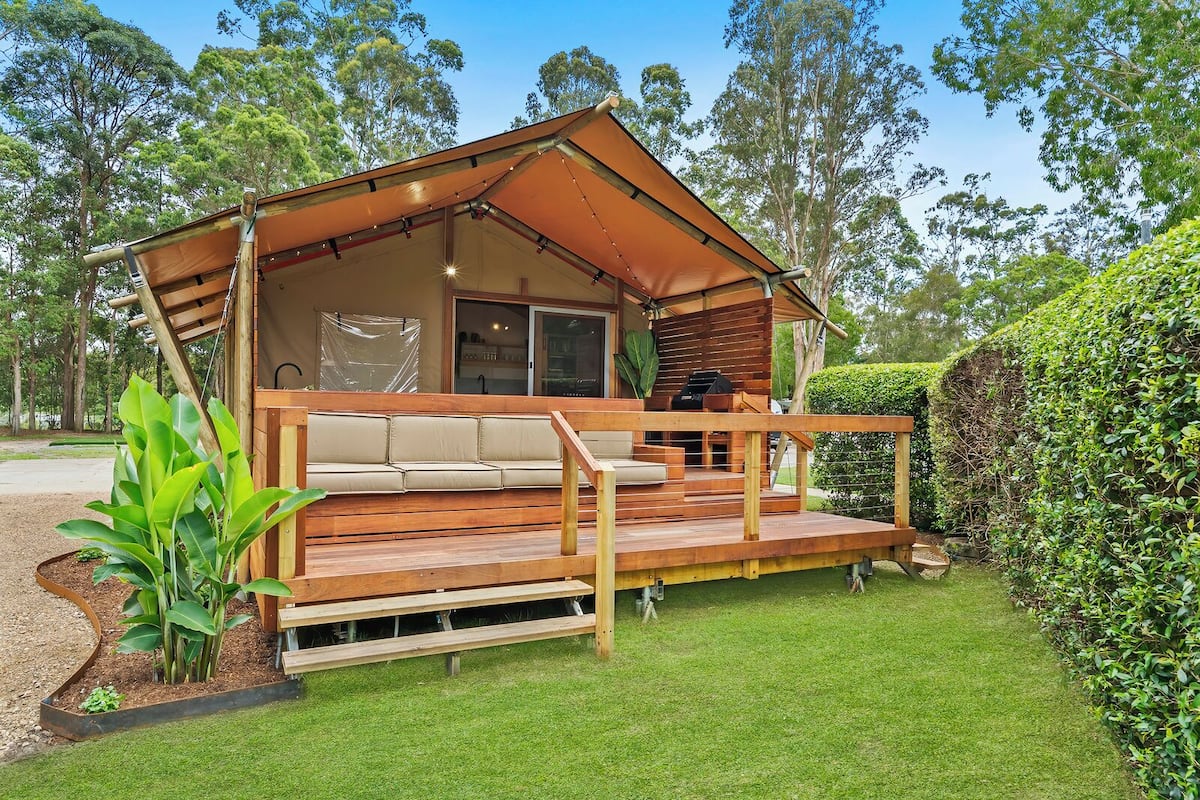 A wooden deck with a sofa and grill area, surrounded by greenery and trees.