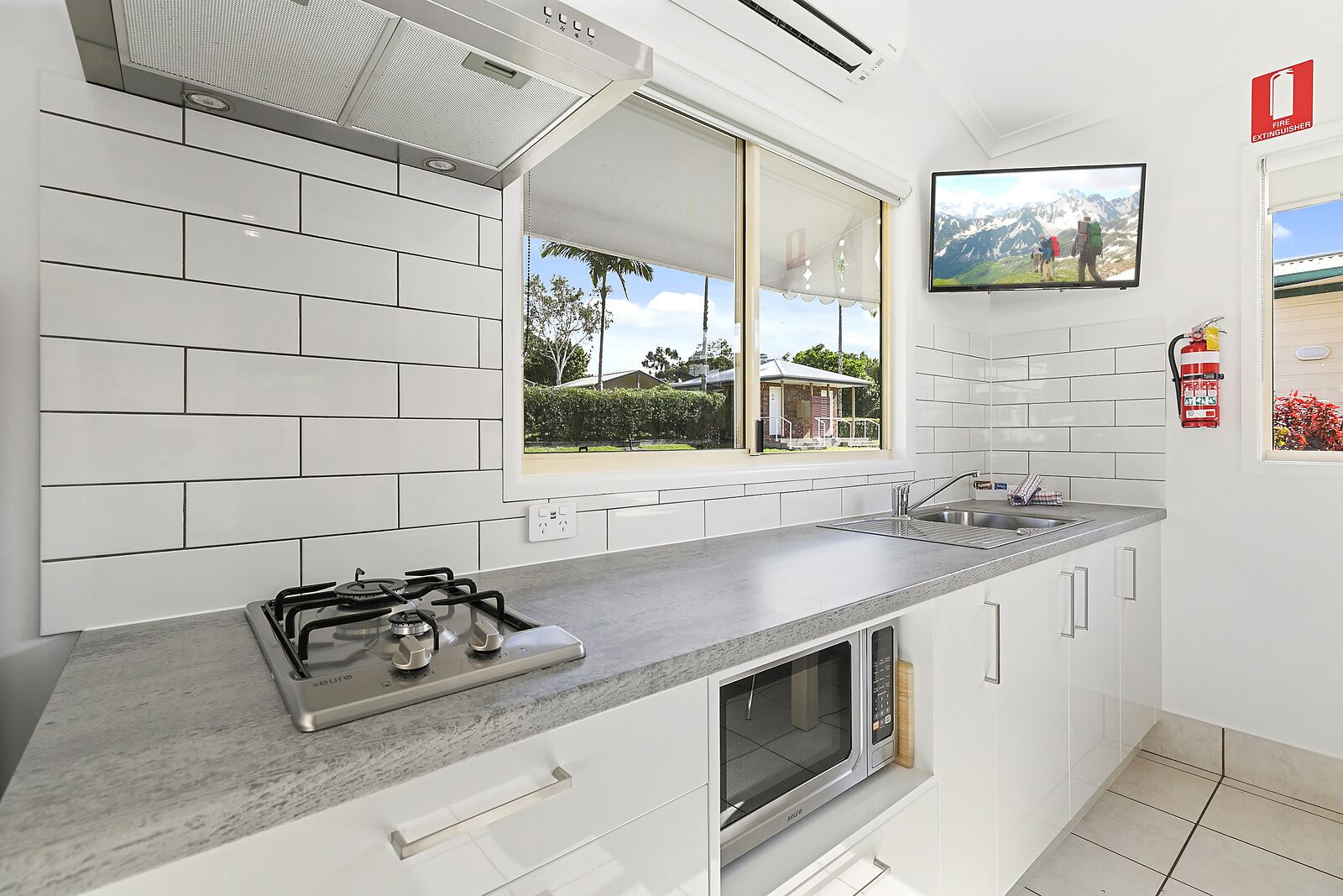 Una cocina moderna con armarios blancos, estufa a gas, microondas y un amplio ventanal con vistas a una casa y follaje.