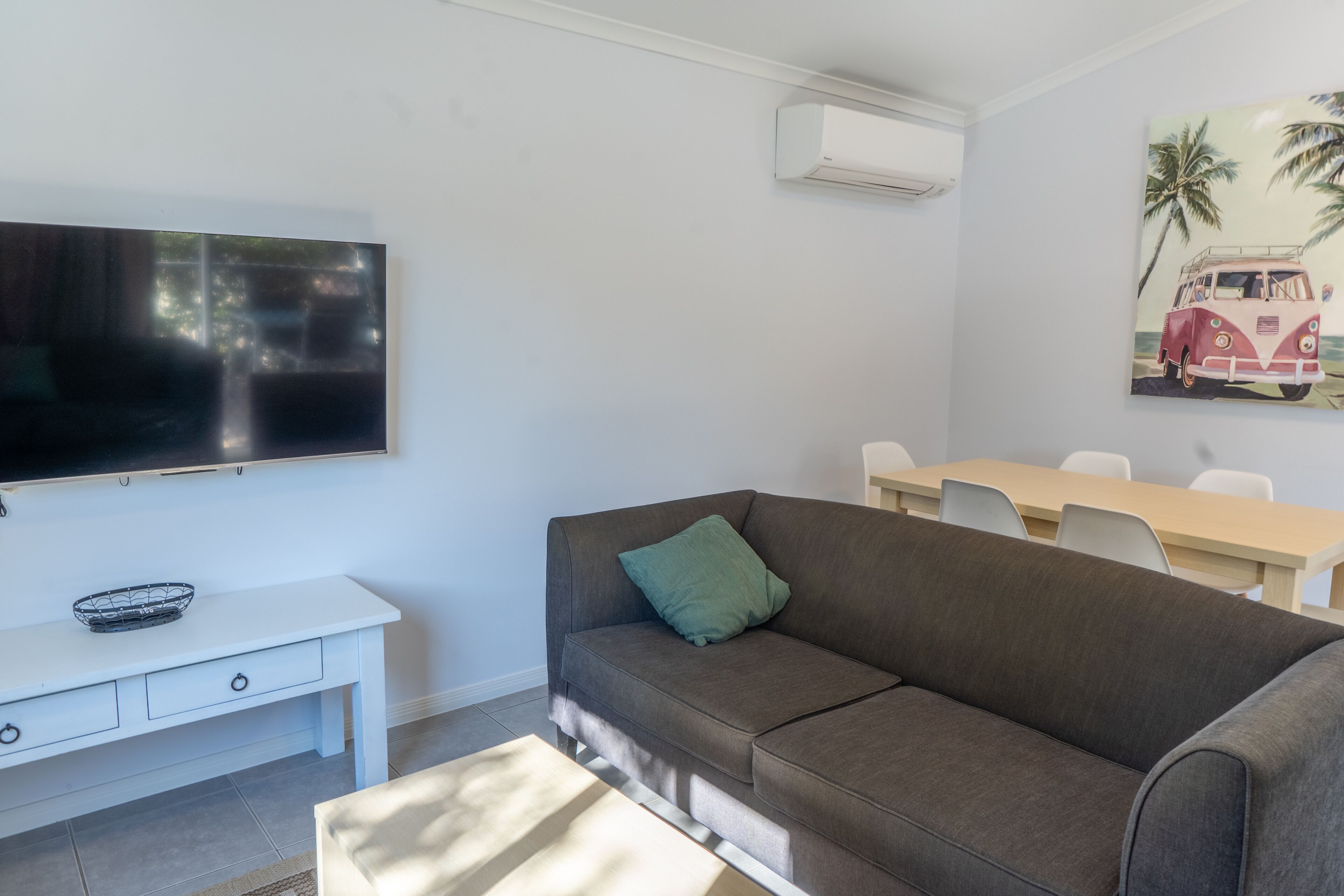 A living room with a flat-screen TV, a gray sofa, a white coffee table, a white console table with a basket, and a painting of a van on the wall.