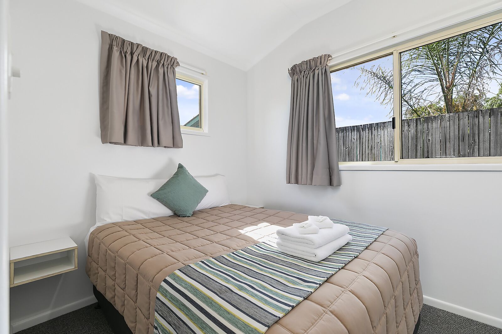 A bedroom with a bed, a window with curtains, a bedside table, and a view of a wooden fence outside.