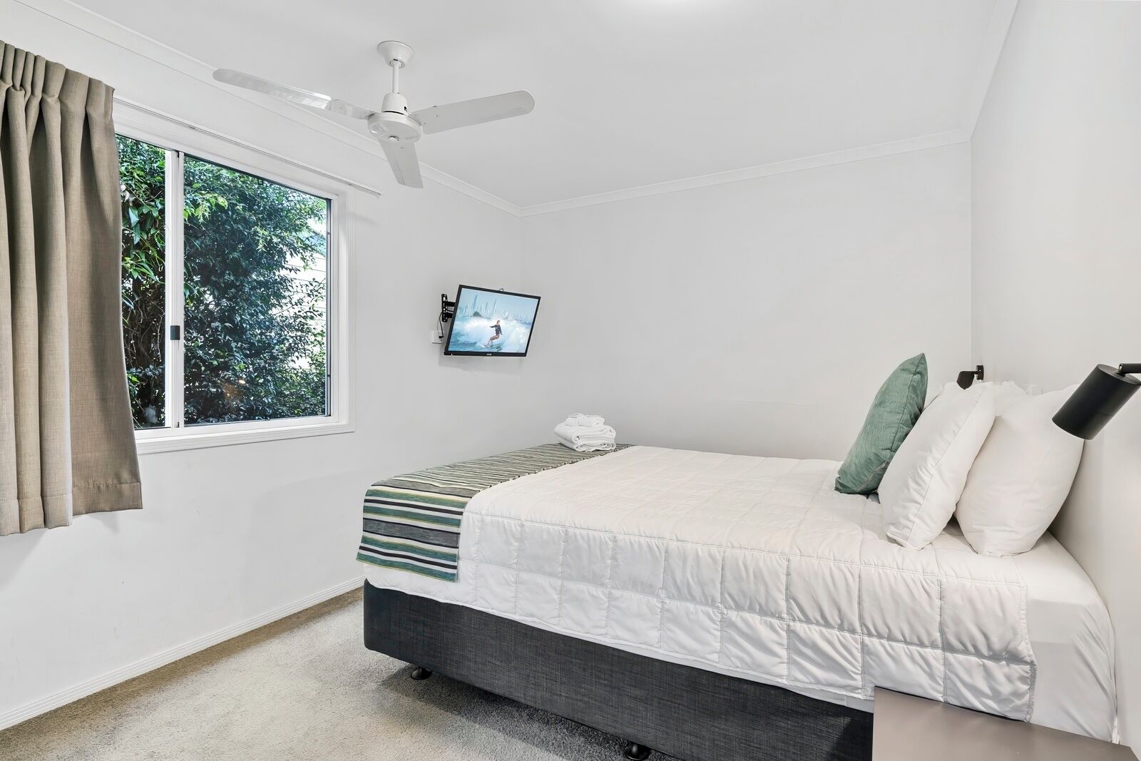 A bedroom with a bed, a ceiling fan, a TV, and a window with curtains.