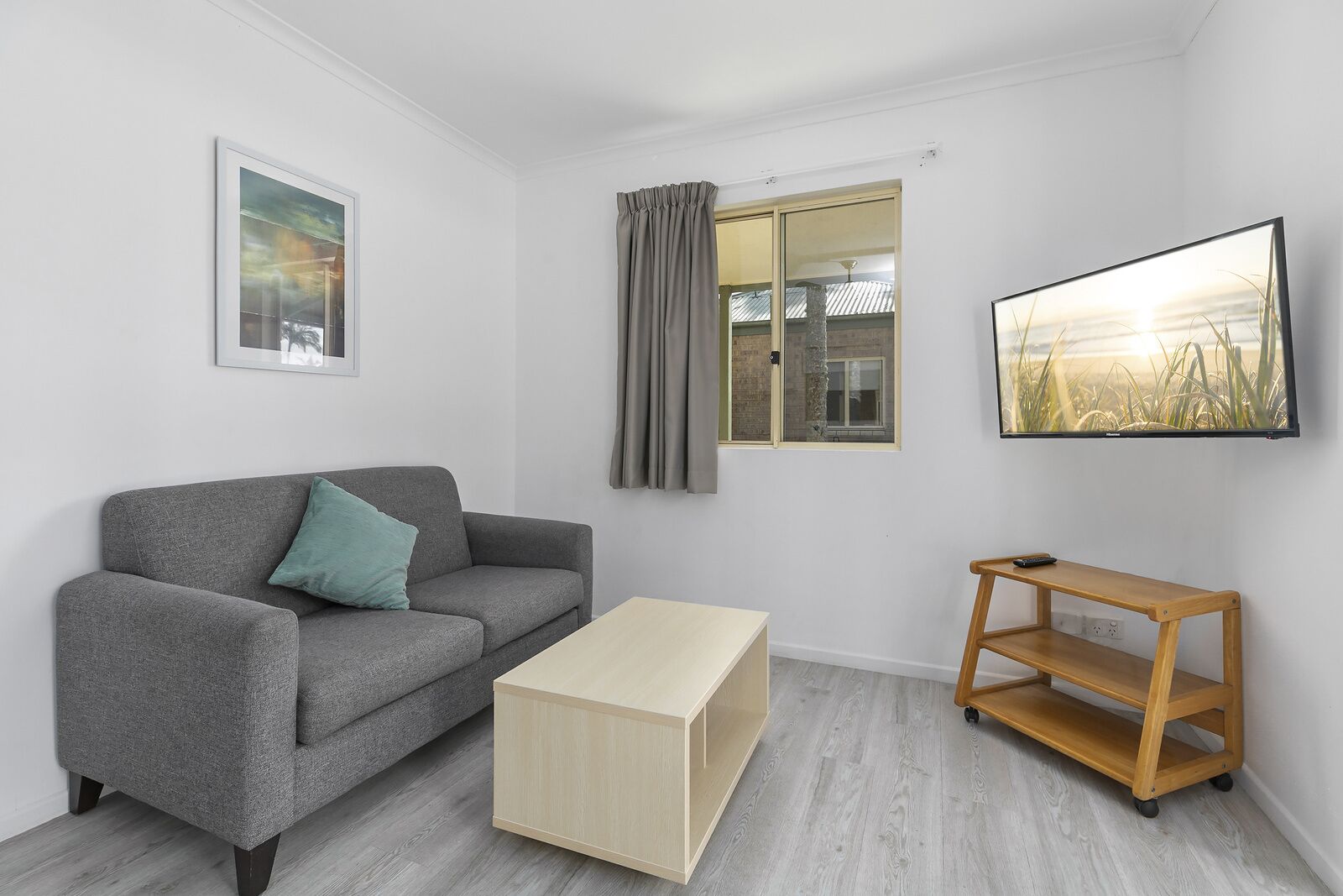 Una sala de estar moderna con un sofá gris, una mesa de centro de madera, un televisor en la pared y un ventanal con cortinas.