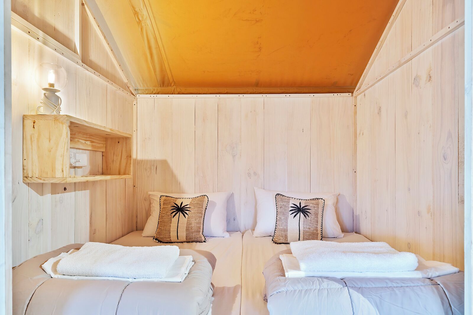 A cozy room with two beds, each with a white blanket and a palm tree pillow. There is a wooden shelf with a lamp and a power outlet.