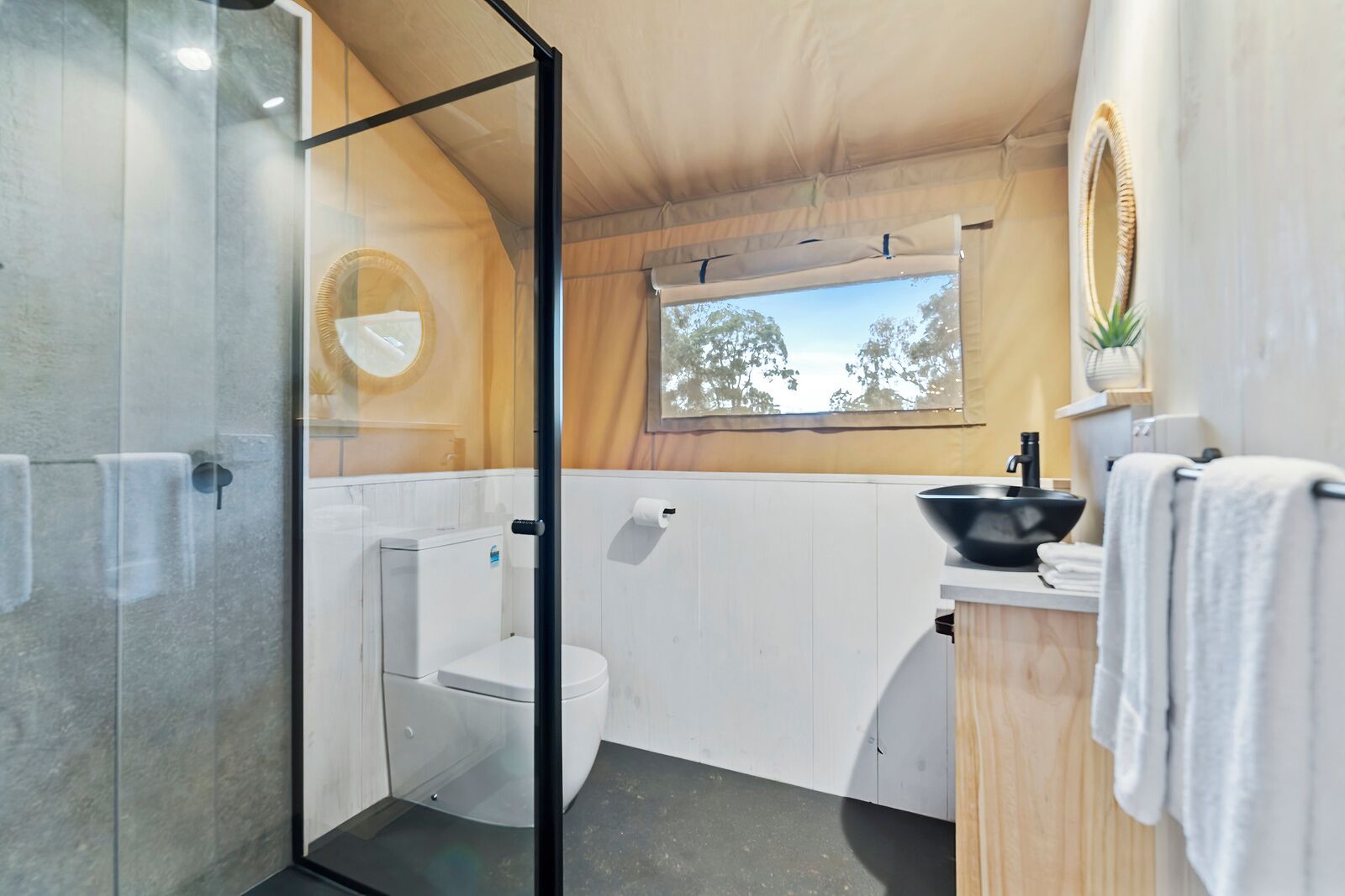 A compact bathroom with a glass shower enclosure, a white toilet, a sink with a black basin, and a window with a view of trees.