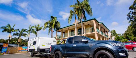 A blue pickup truck towing a white camper trailer with "EXPAND" branding, parked in front of a two-story building with balconies and palm trees in the background.