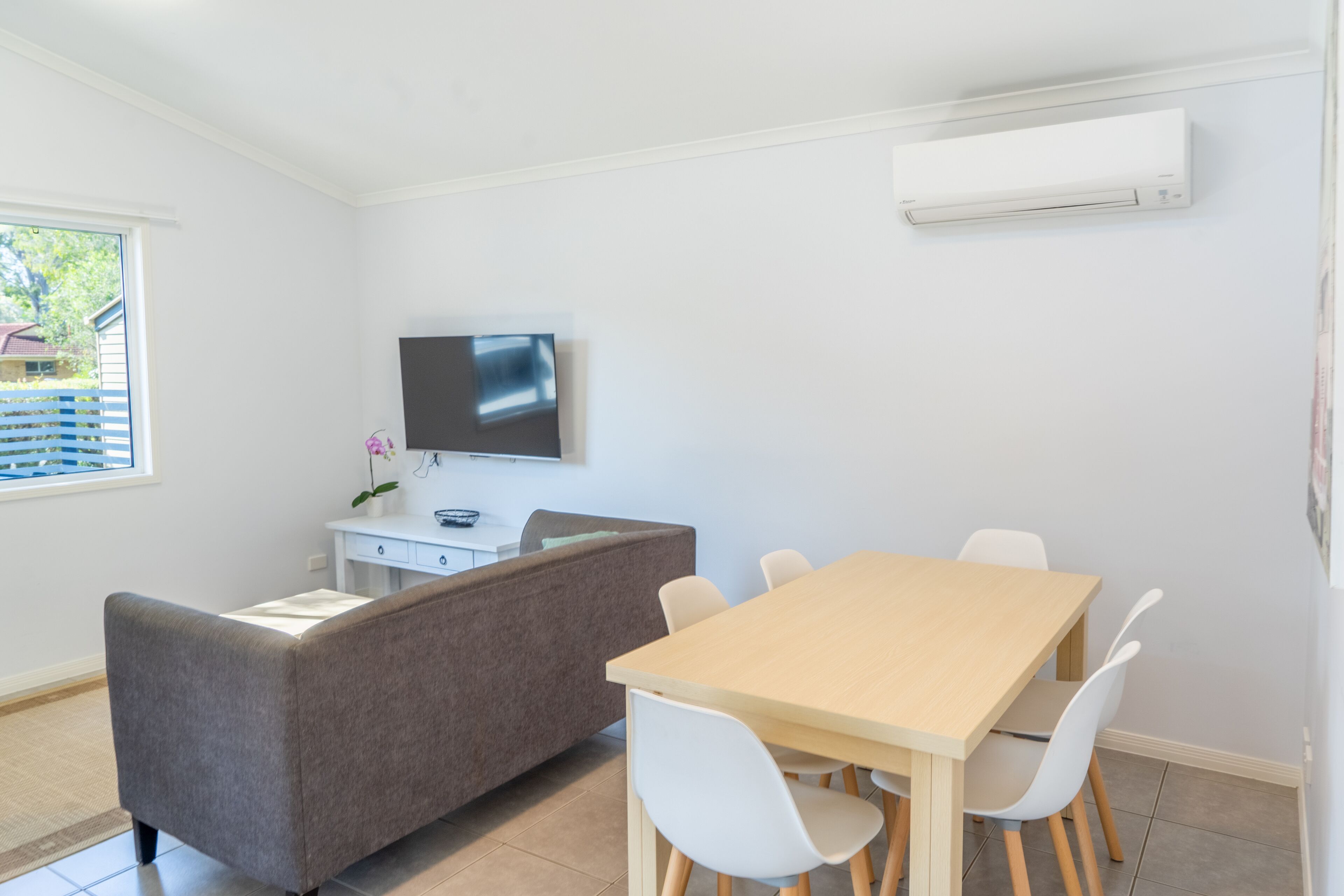 Une pièce moderne avec une table à manger en bois, des chaises blanches, un canapé gris, un téléviseur à écran plat fixé au mur et un petit bureau blanc agrémenté d’une plante.