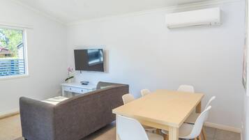 Une pièce moderne avec une table à manger en bois, des chaises blanches, un canapé gris, un téléviseur à écran plat fixé au mur et un petit bureau blanc agrémenté d’une plante.