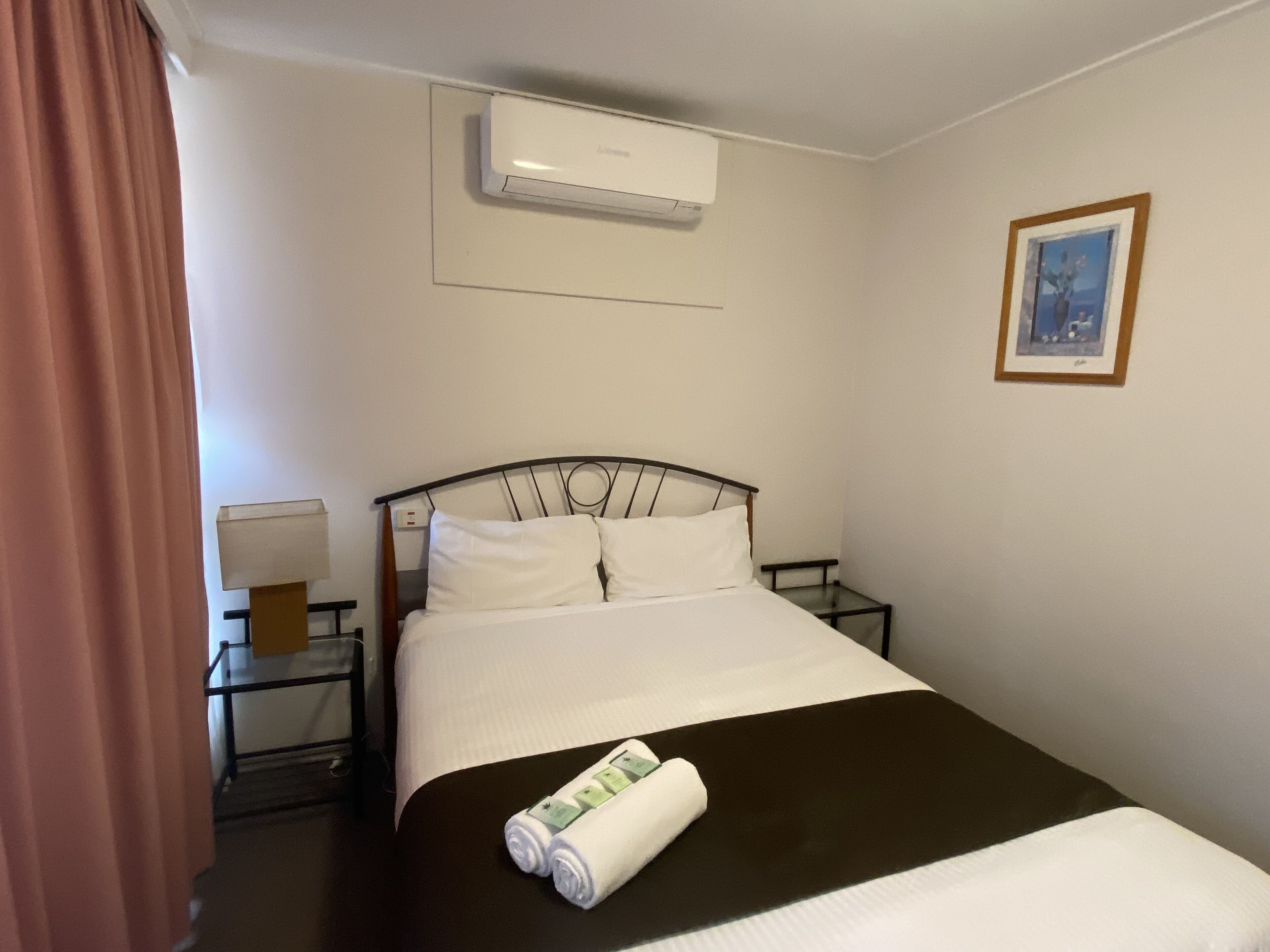 Une chambre d’hôtel avec un lit, une table de chevet, un appareil de climatisation, un tableau encadré au mur et un rideau.