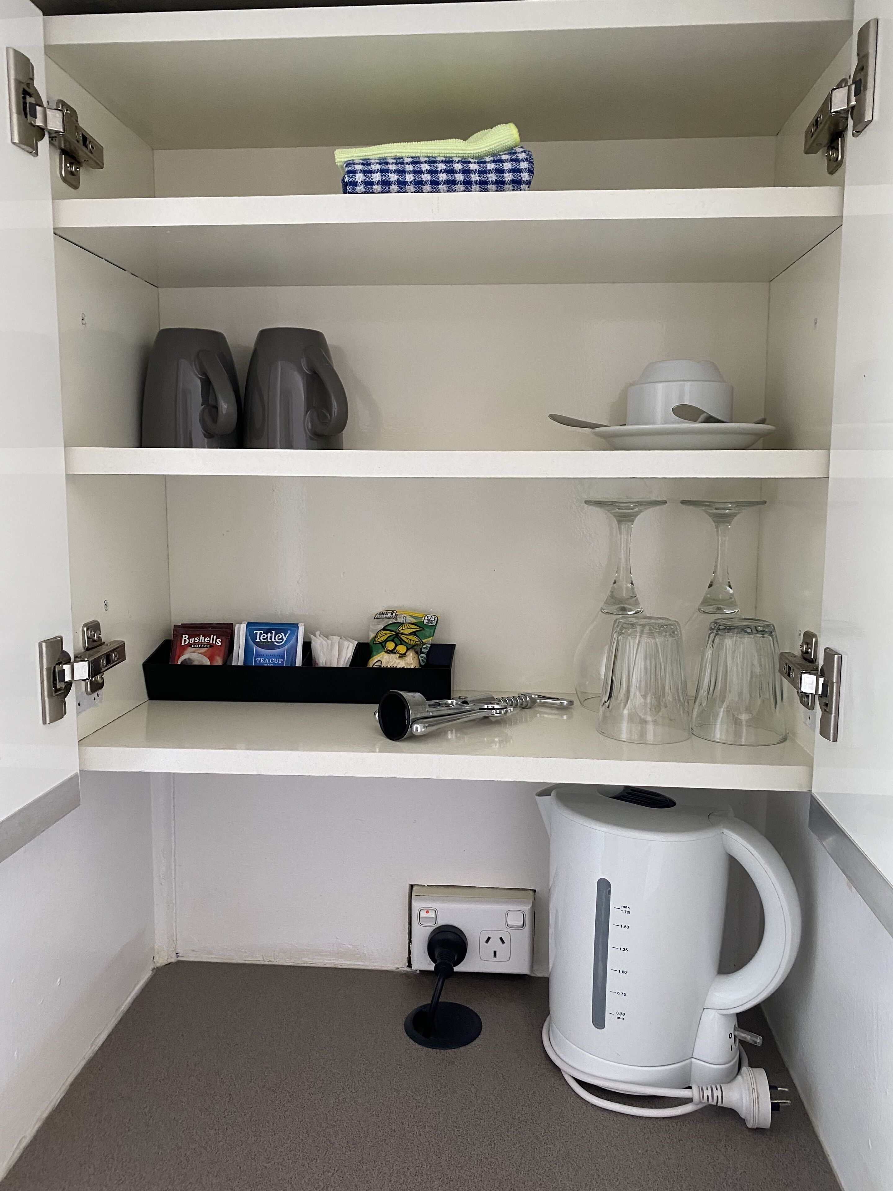 A compact kitchenette with a coffee maker, two mugs, a tea set, and a small shelf with condiments.