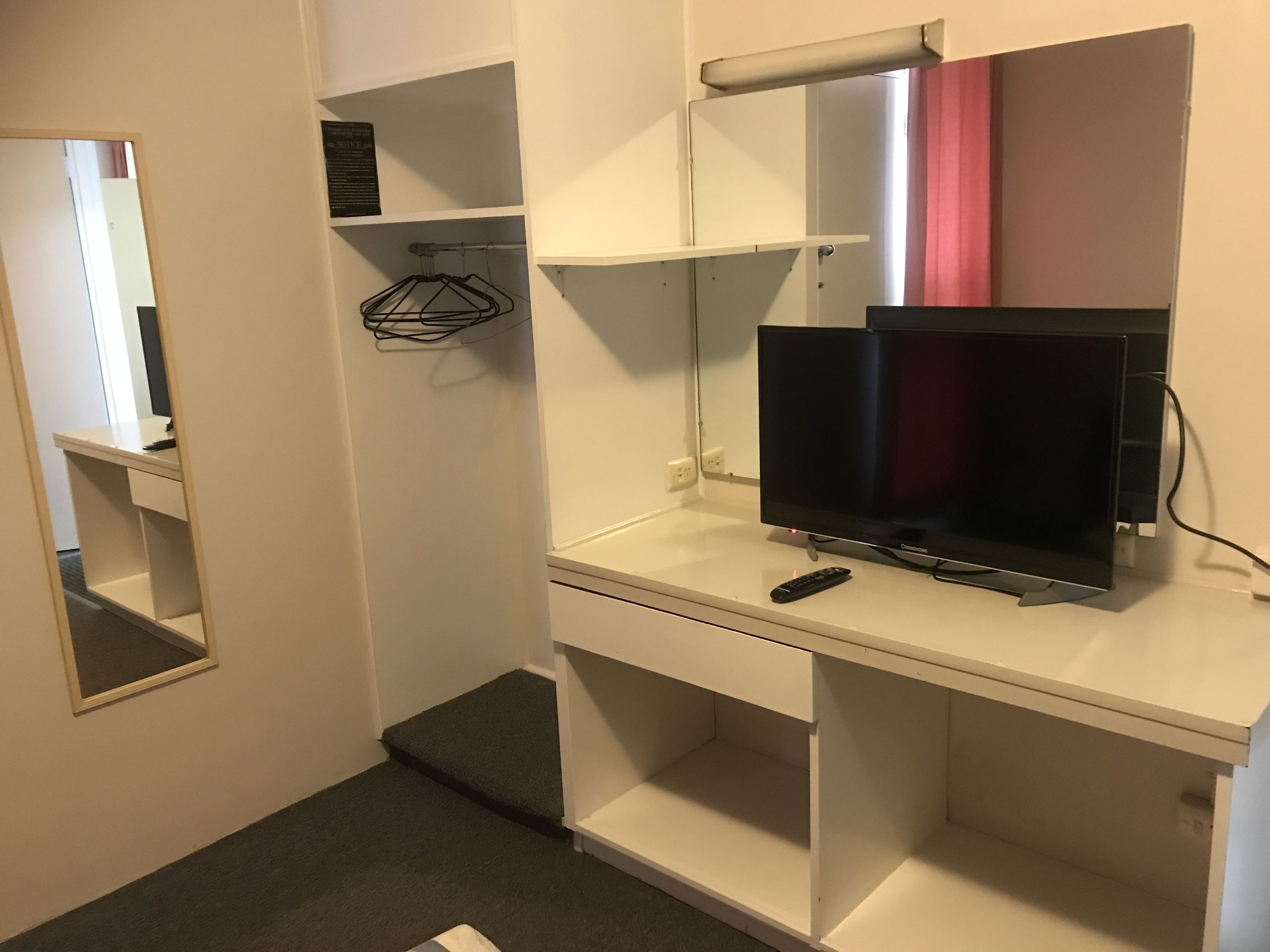 Habitación de hotel con televisor sobre un soporte blanco, un espejo y un estante con perchero.