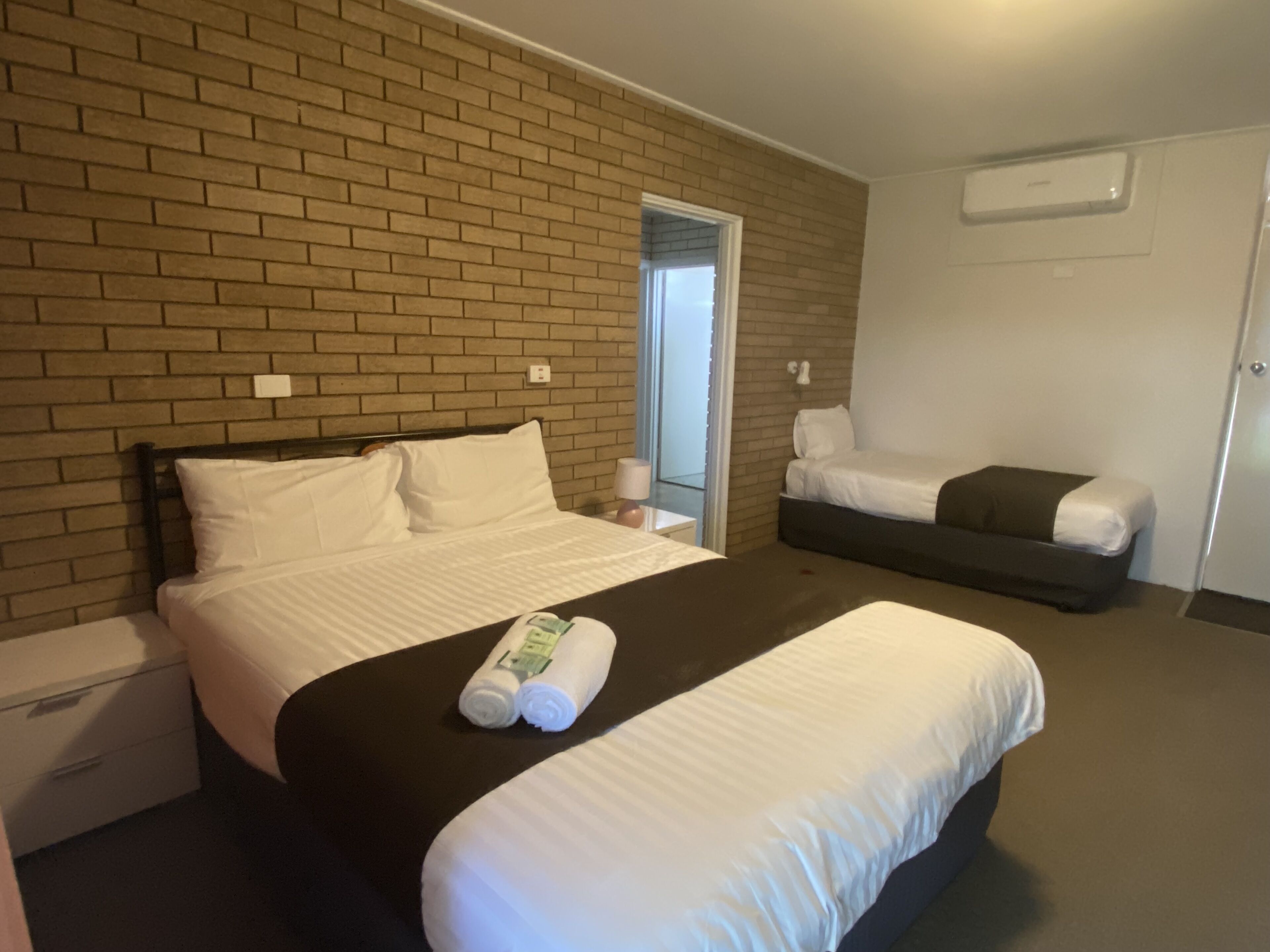 A hotel room with a brick wall, a large bed with white bedding, a smaller bed, a nightstand, and a door.