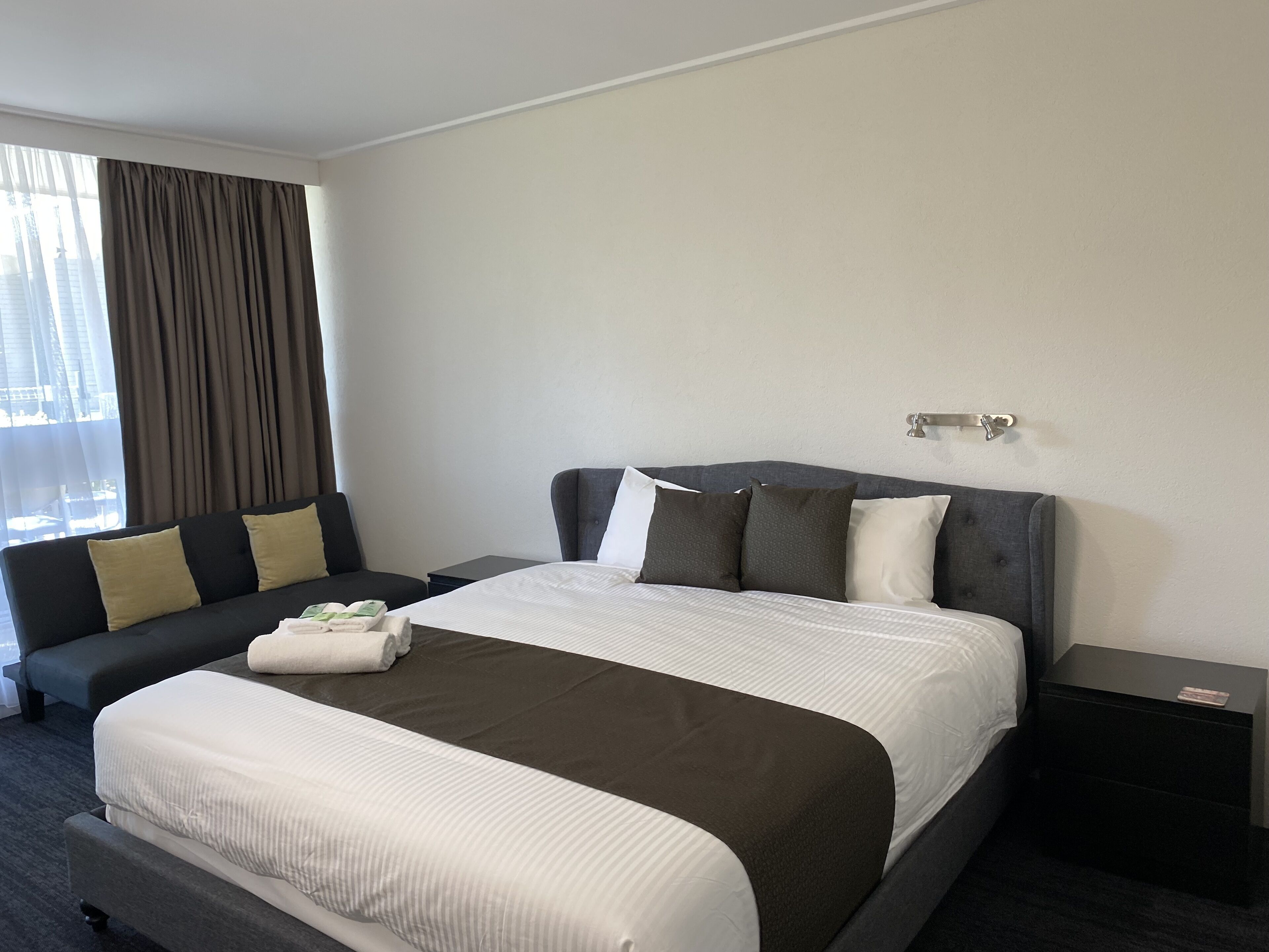 A modern hotel room with a large bed, a sofa, a nightstand, and a window with curtains.