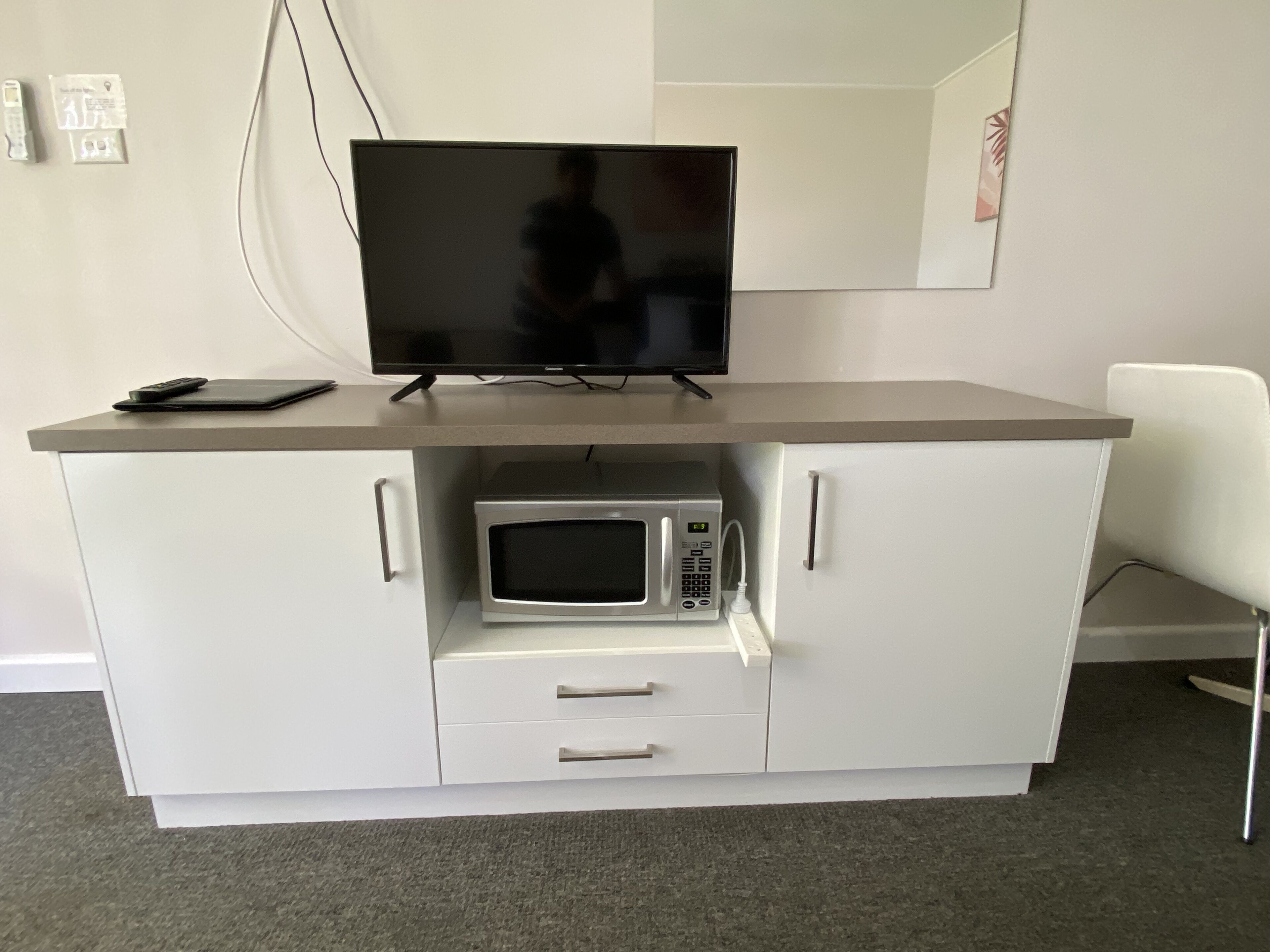 A kitchenette with a microwave, a small fridge, a flat-screen TV, and a white chair.