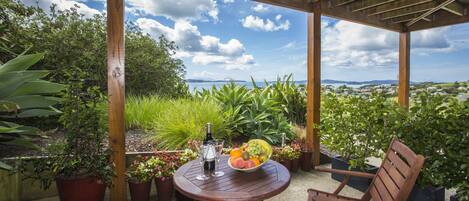 A wooden pergola with a table and chairs, a bowl of fruit, and a bottle of wine.