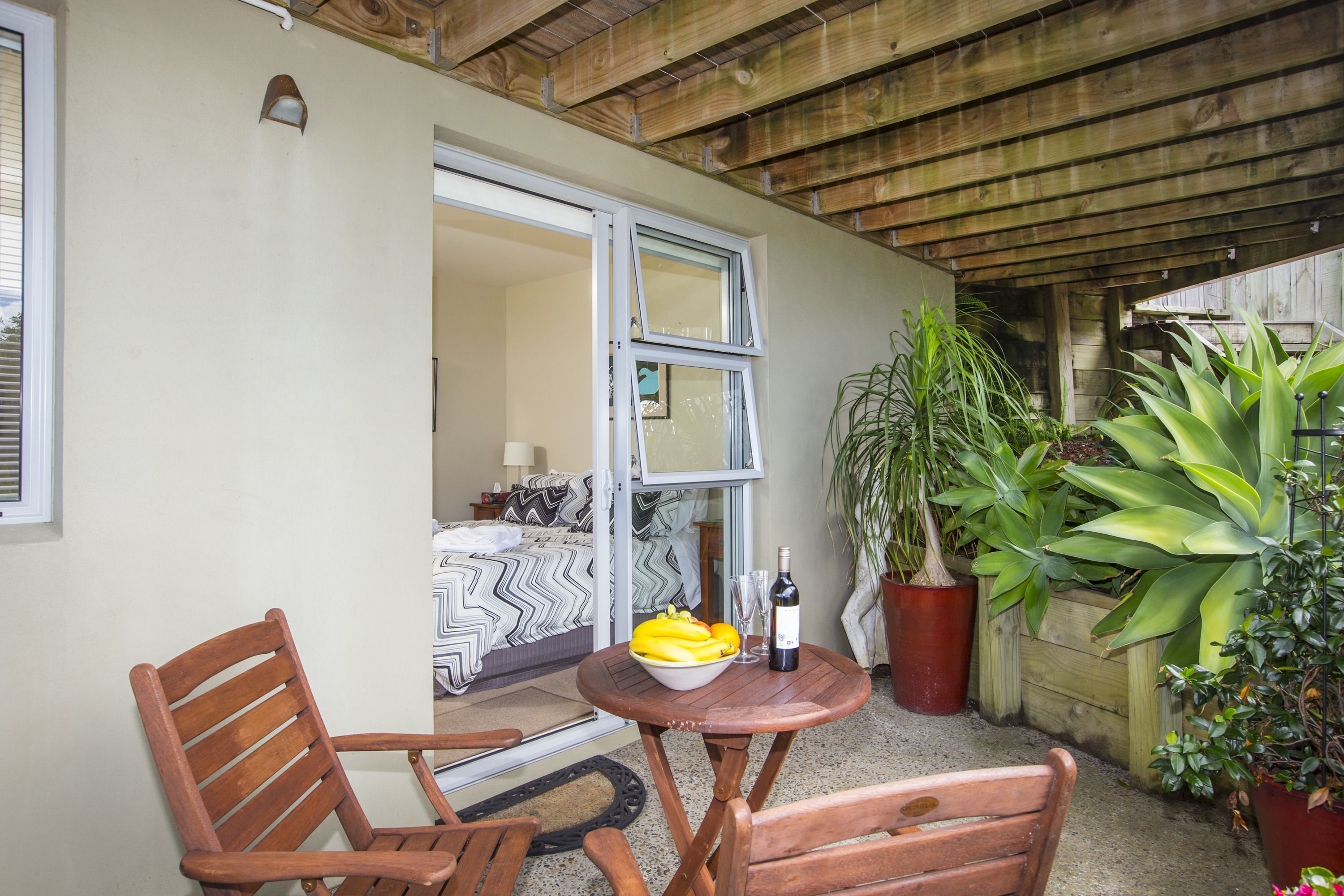 A patio with wooden furniture, a table with a bowl of fruit and a bottle, and potted plants.