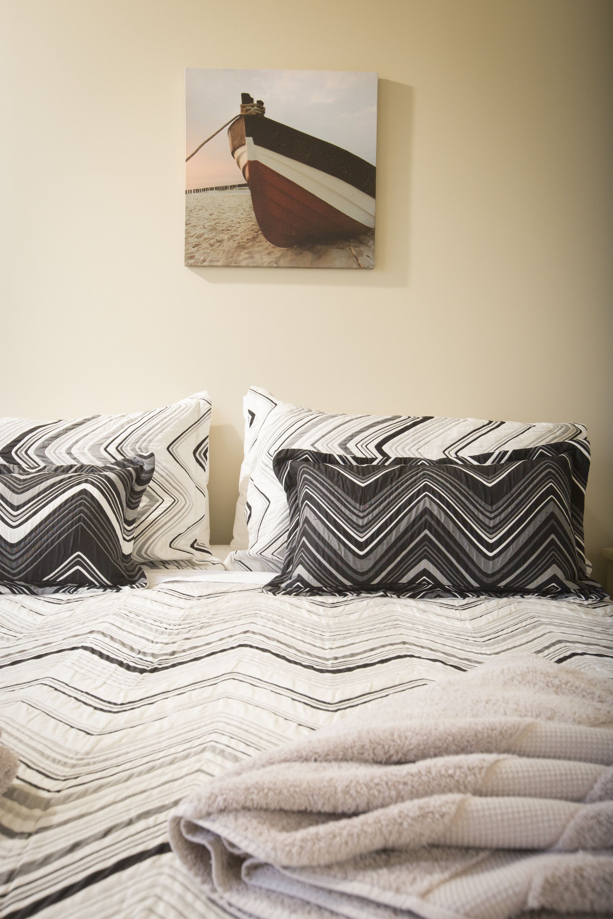 A bed with a patterned bedspread, two pillows with a zigzag design, and a painting of a boat on the wall.