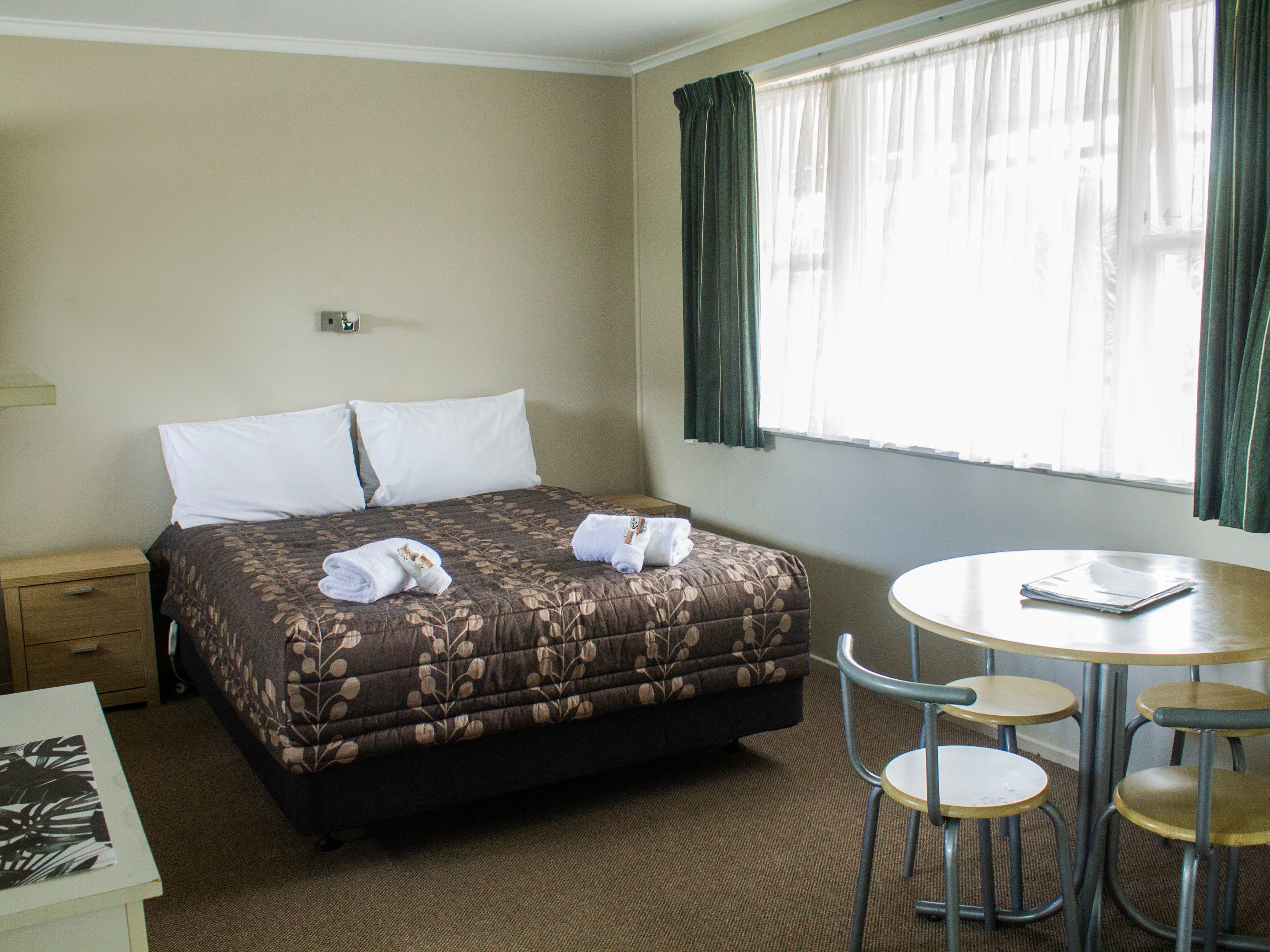A hotel room with a bed, a small table with chairs, a nightstand, and a window with curtains.