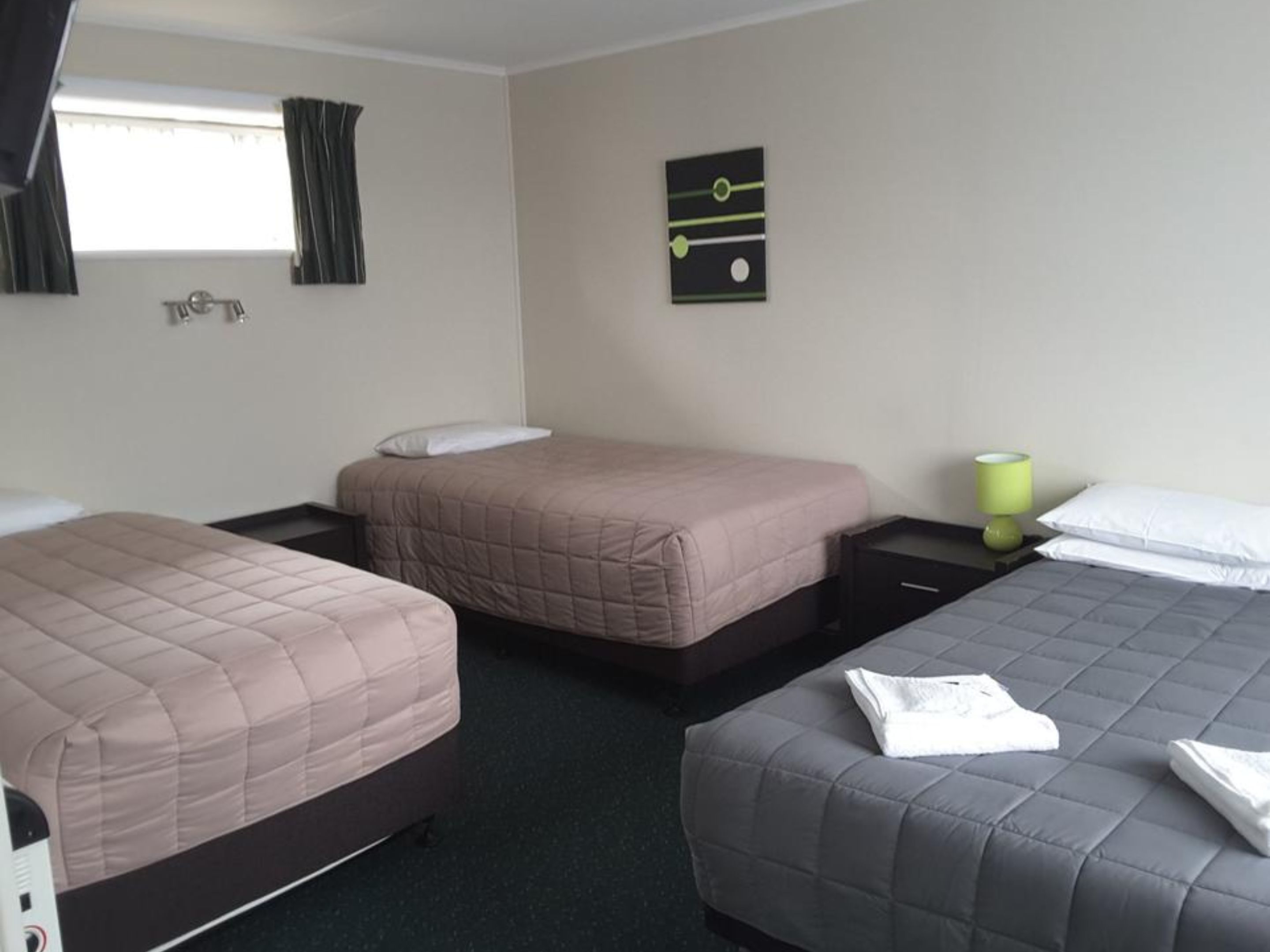 A hotel room with three beds, a small table, a lamp, and a wall clock.