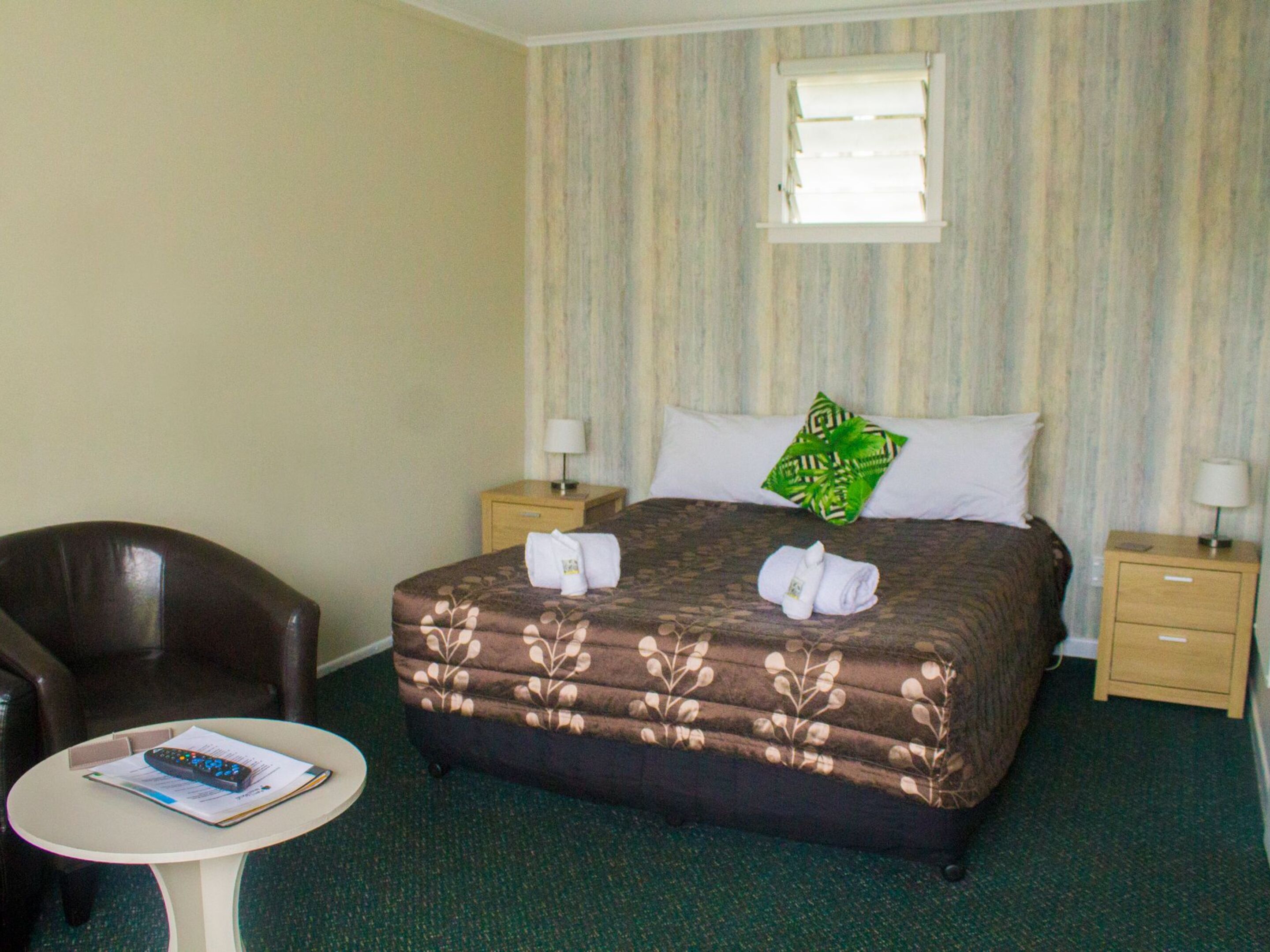 Quarto de hotel com uma cama, duas mesas de cabeceira, uma cadeira e uma mesinha redonda com um controle remoto e um livreto.