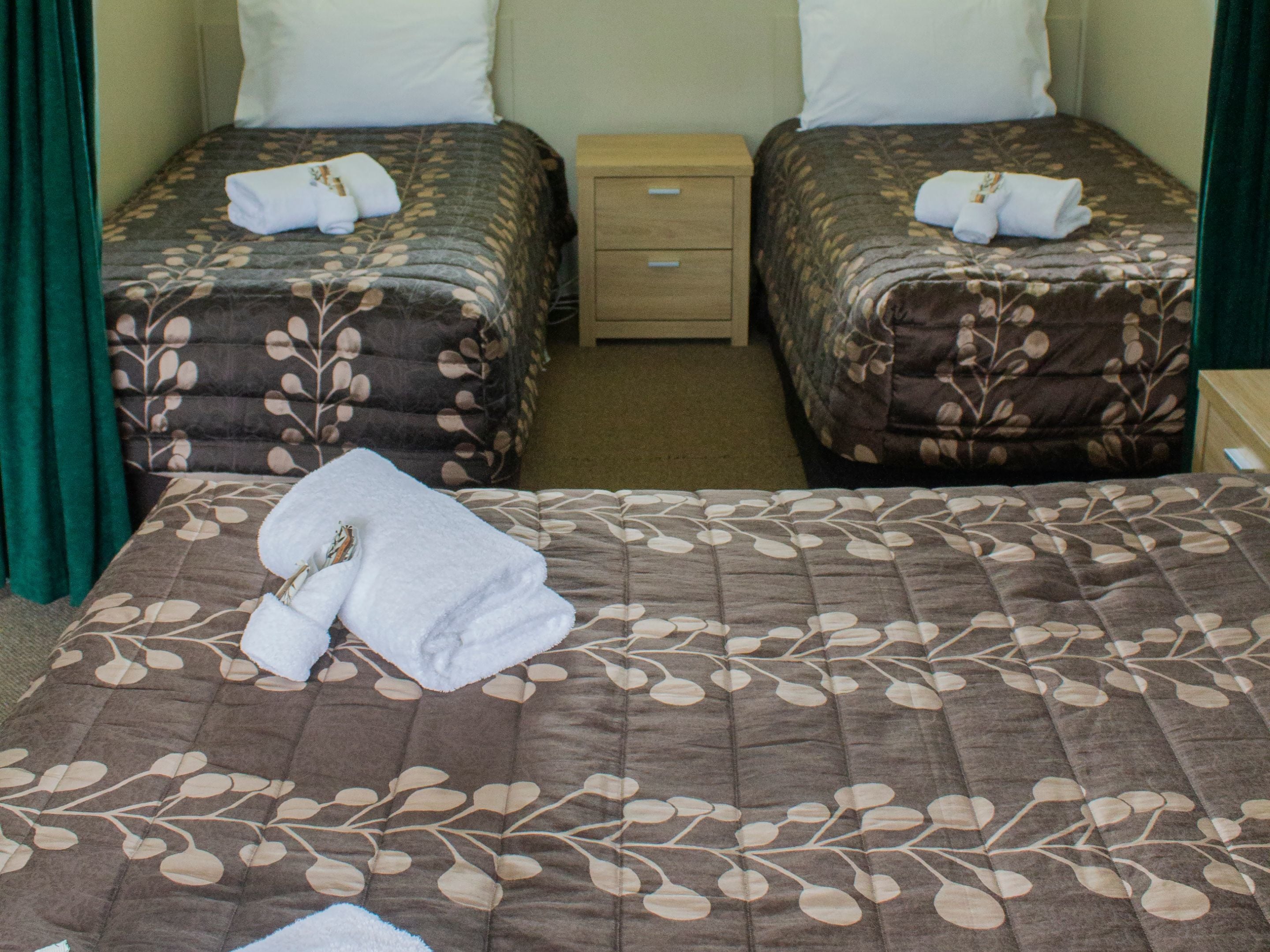 A hotel room with two single beds, each with a floral-patterned bedspread and a folded towel. There is a wooden nightstand between the beds.