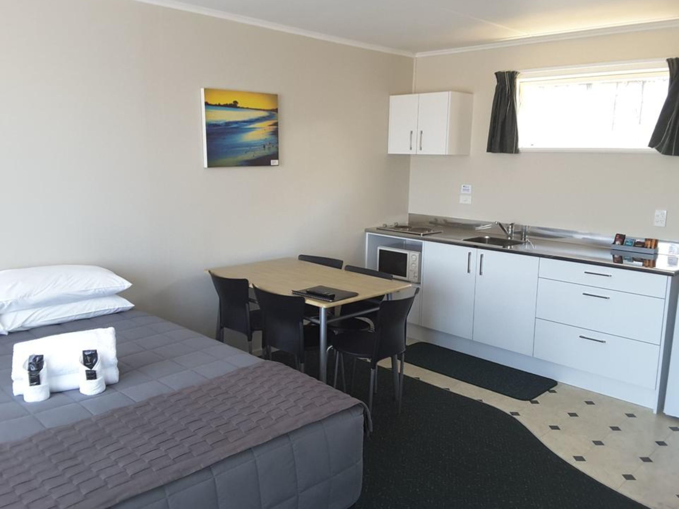 A compact hotel room with a bed, a small dining table, chairs, a kitchenette, and a painting on the wall.