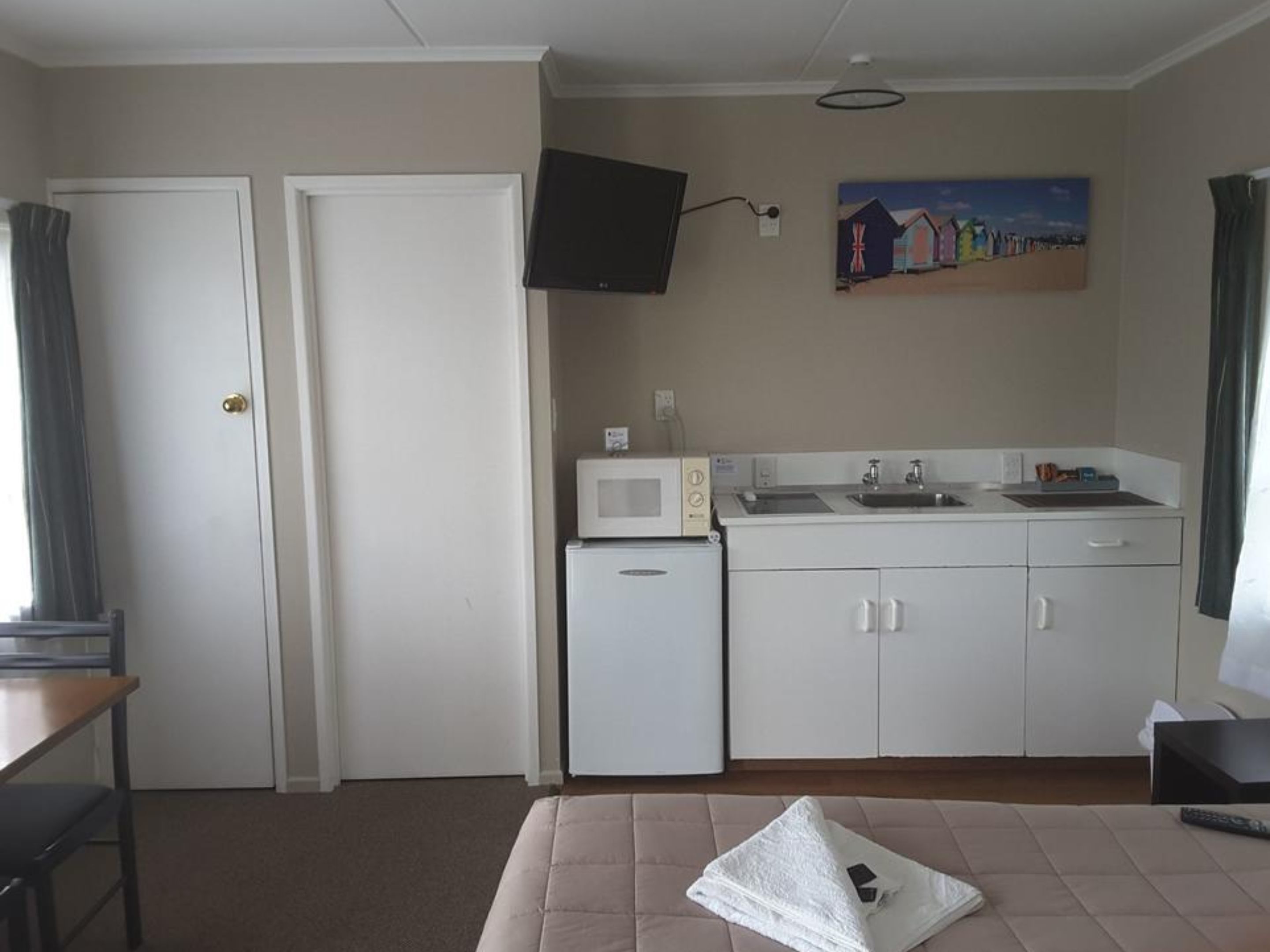 A compact hotel room with a kitchenette, a bed, a desk, and a painting on the wall.