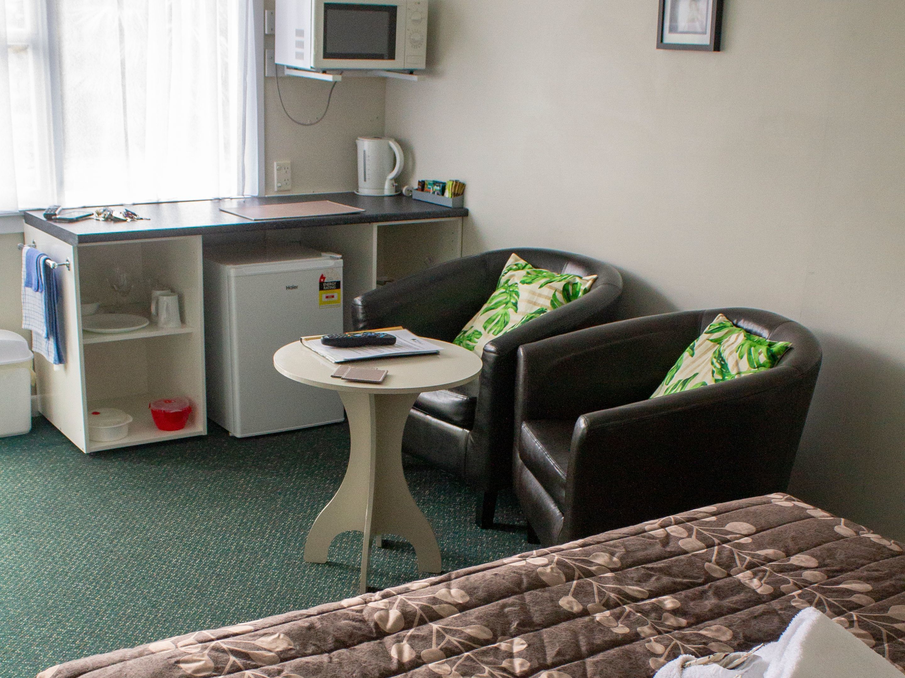 A compact hotel room with a bed, a small desk with a microwave, a coffee maker, and a chair with a pillow.