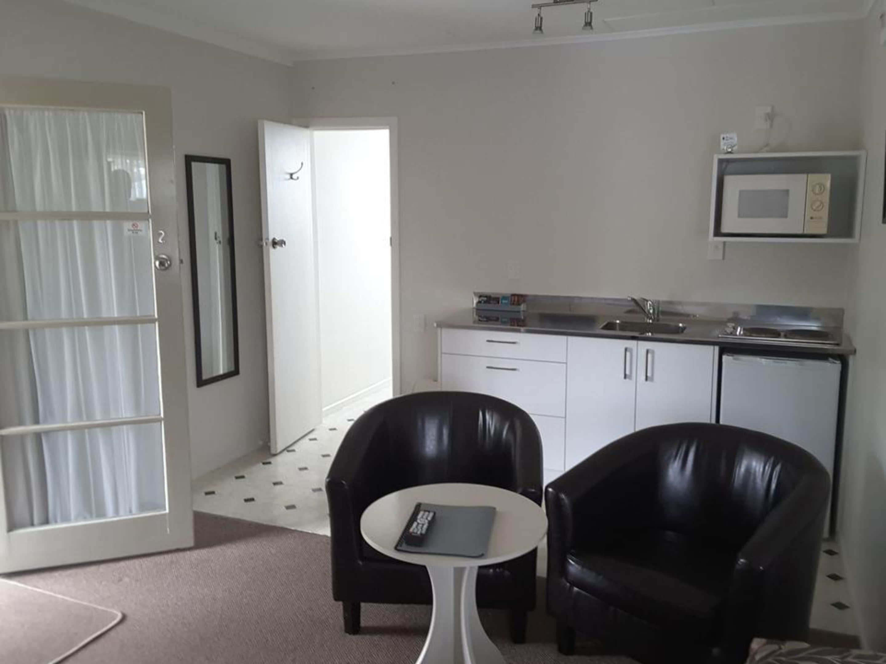 A compact room with a kitchenette, two black leather chairs, a small white table, and a door leading to another room.